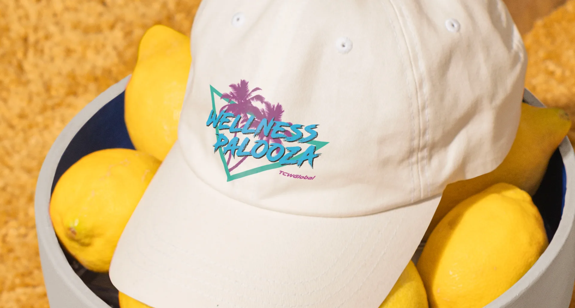 White baseball cap embroidered with the pink and teal Wellness Palooza palm-tree logo, sitting in a bin of lemons