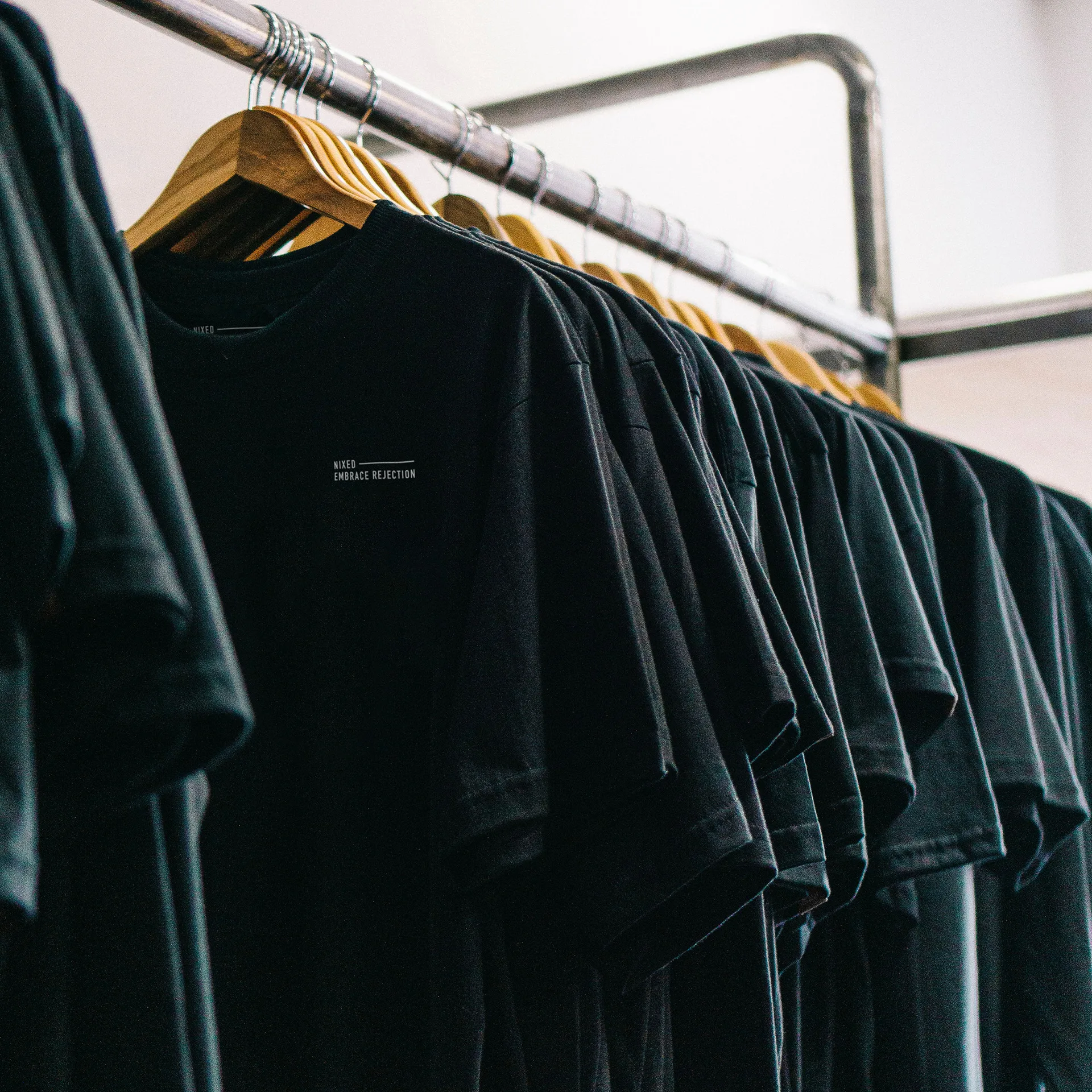 Rack of black NIXED Embrace Rejection t-shirts hanging on wooden hangers in a shop