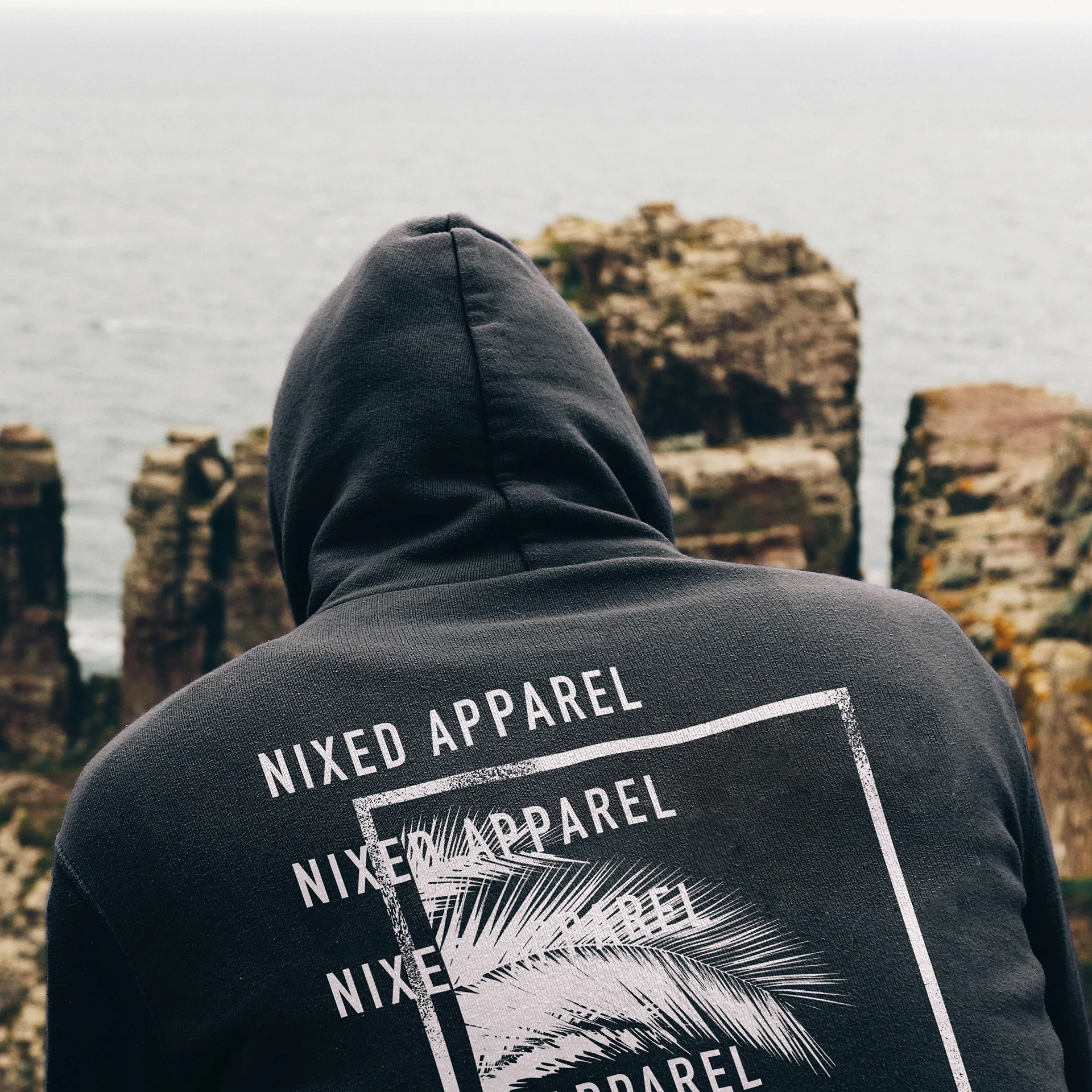 Man's back in a charcoal NIXED Apparel hoodie with repeated wordmark and palm frond print, on coastal cliffs