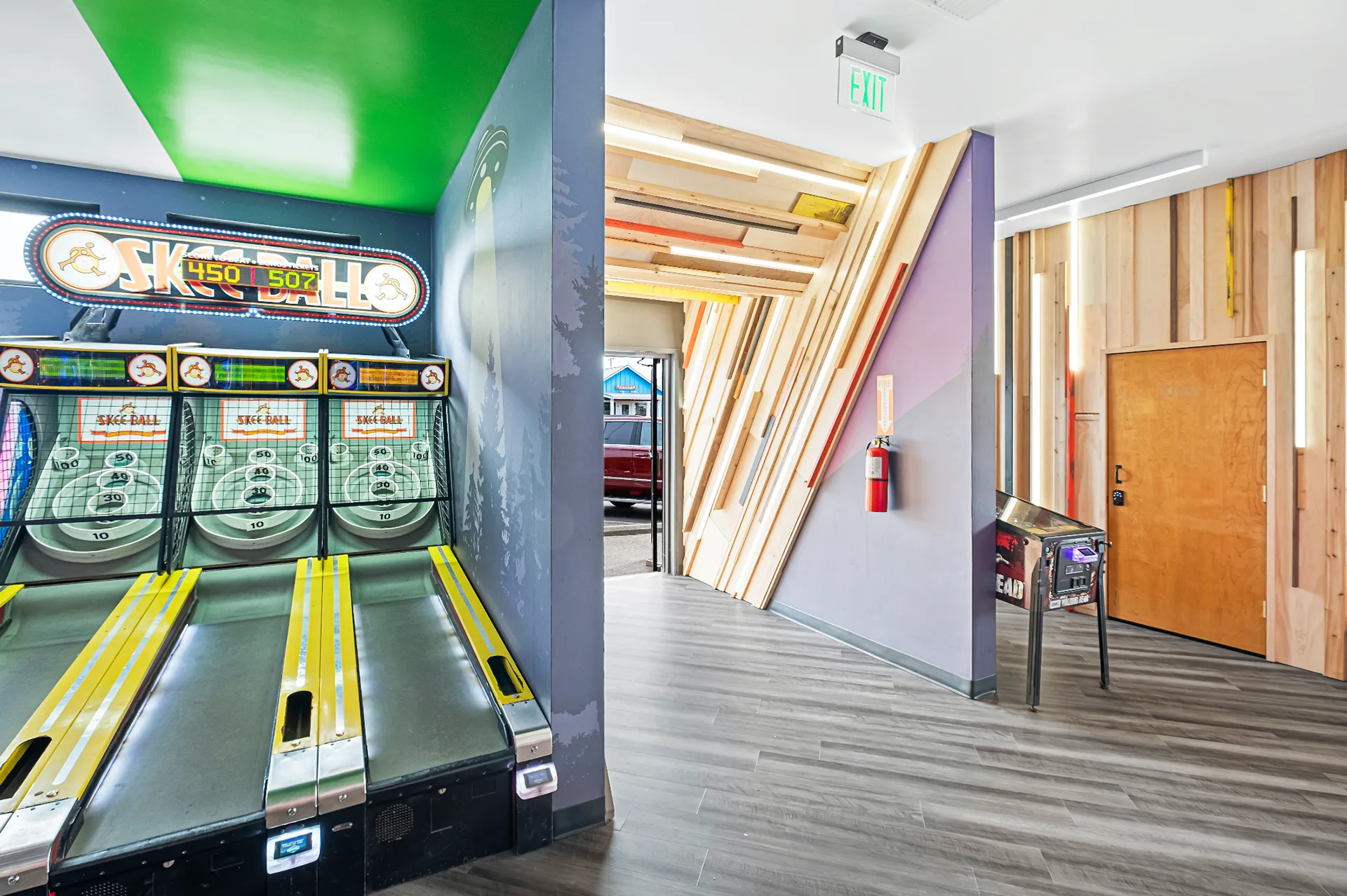 Skee-ball lanes beside a hallway with a raw lumber accent wall leading past an exit sign