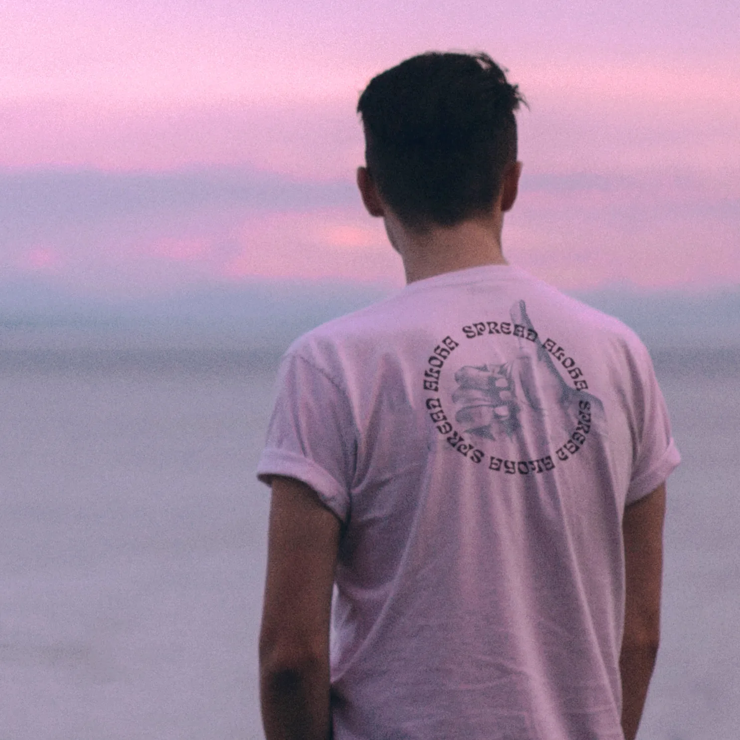Man's back in a pink NIXED tee with a circular Spread Aloha graphic, facing a pastel sunset over the ocean