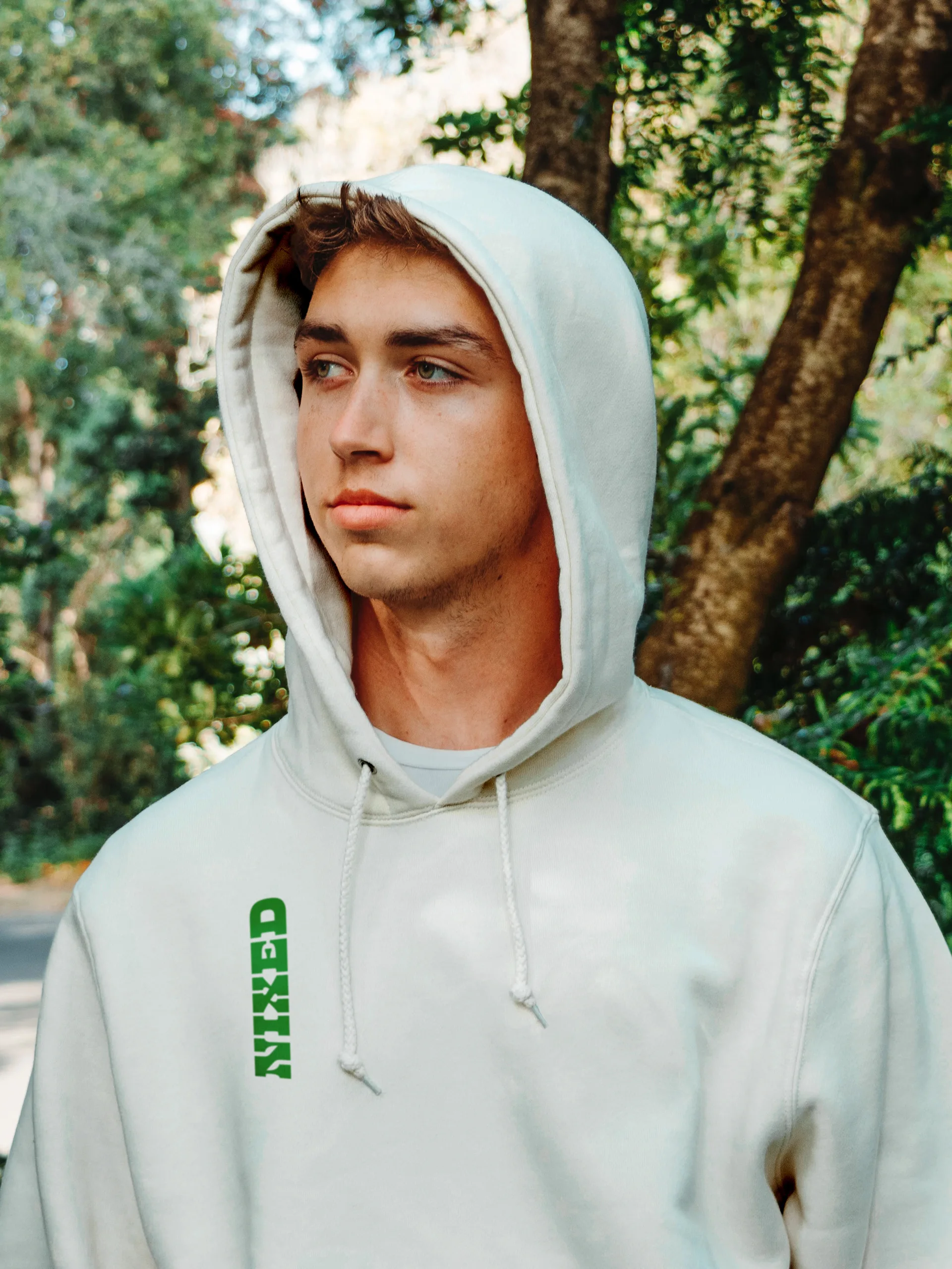 Young man in a cream hoodie with a green vertical NIXED logo down the chest, standing outdoors