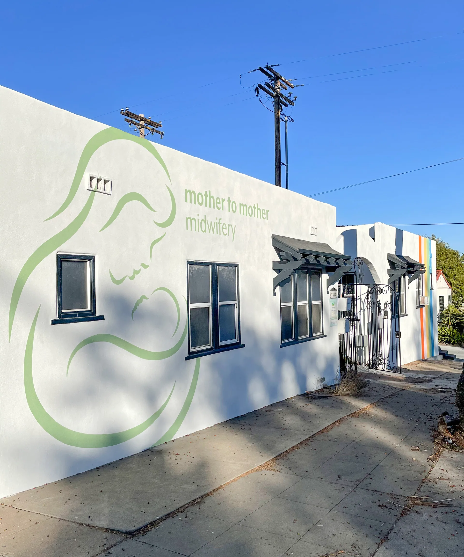 Wide view of the Mother to Mother Midwifery building corner with its logo mural and the rainbow-striped wall beyond