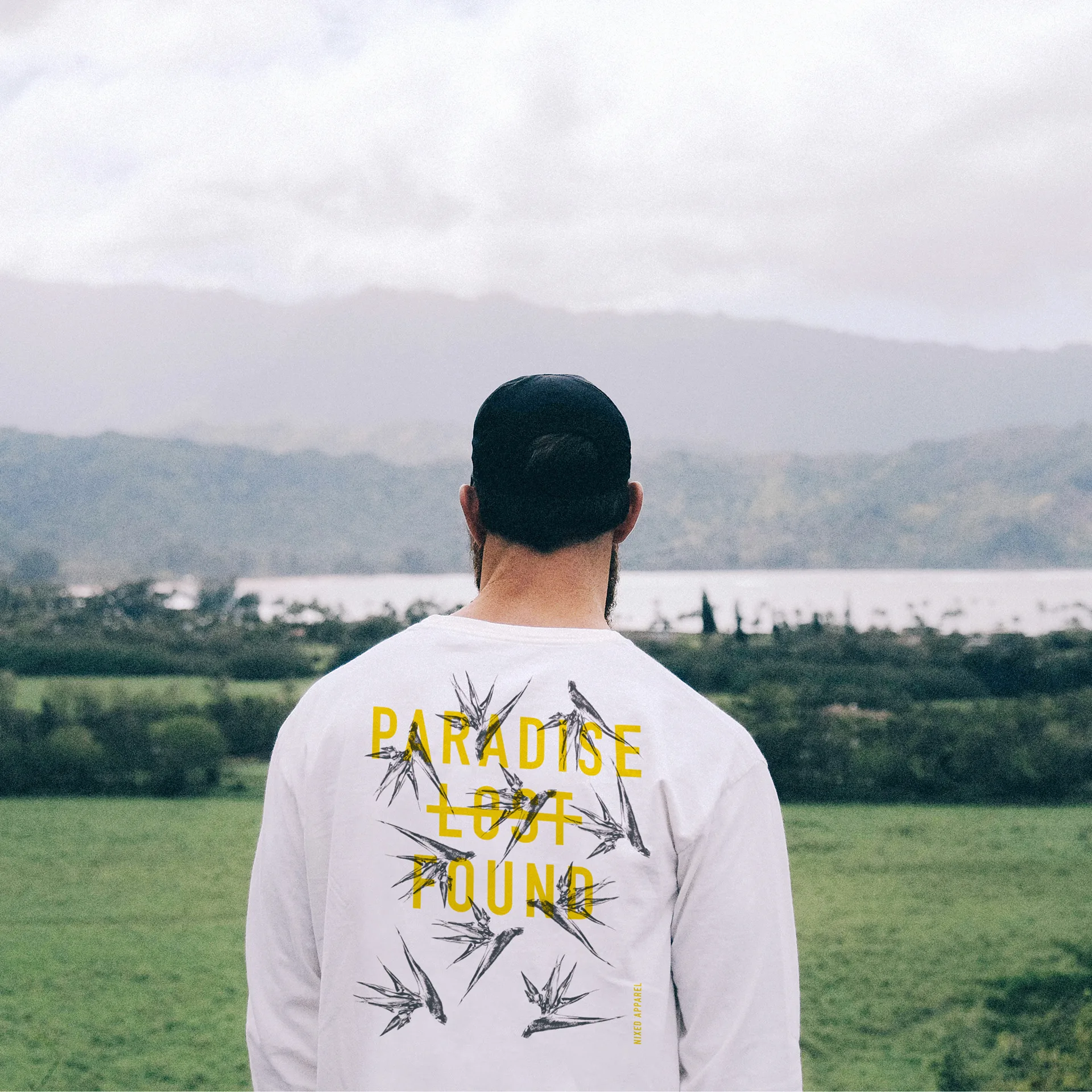 Man in a white long-sleeve NIXED tee printed Paradise Lost Found with bird graphics, facing a green valley