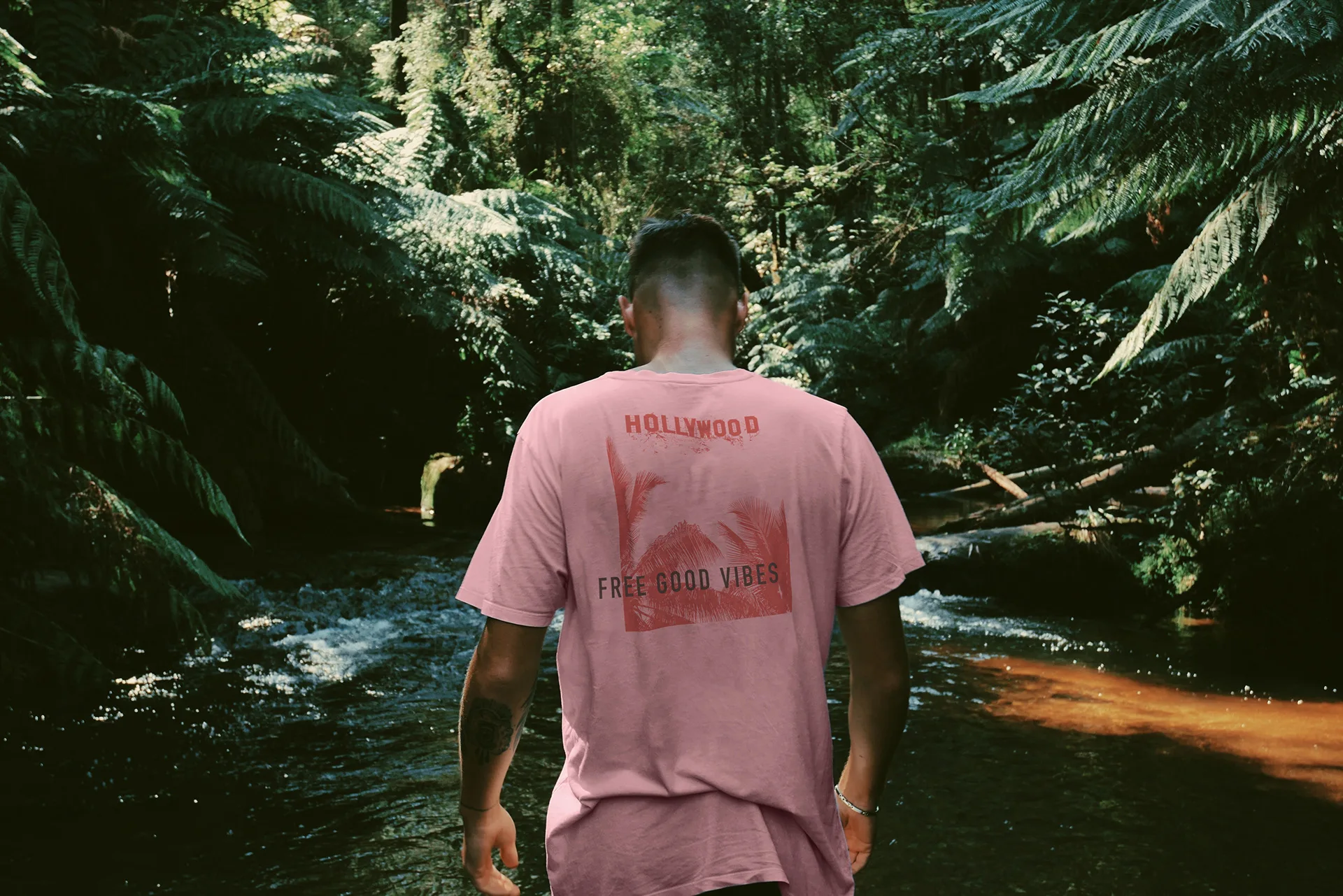 Man's back in a pink NIXED tee reading Hollywood Free Good Vibes, walking through a forest stream