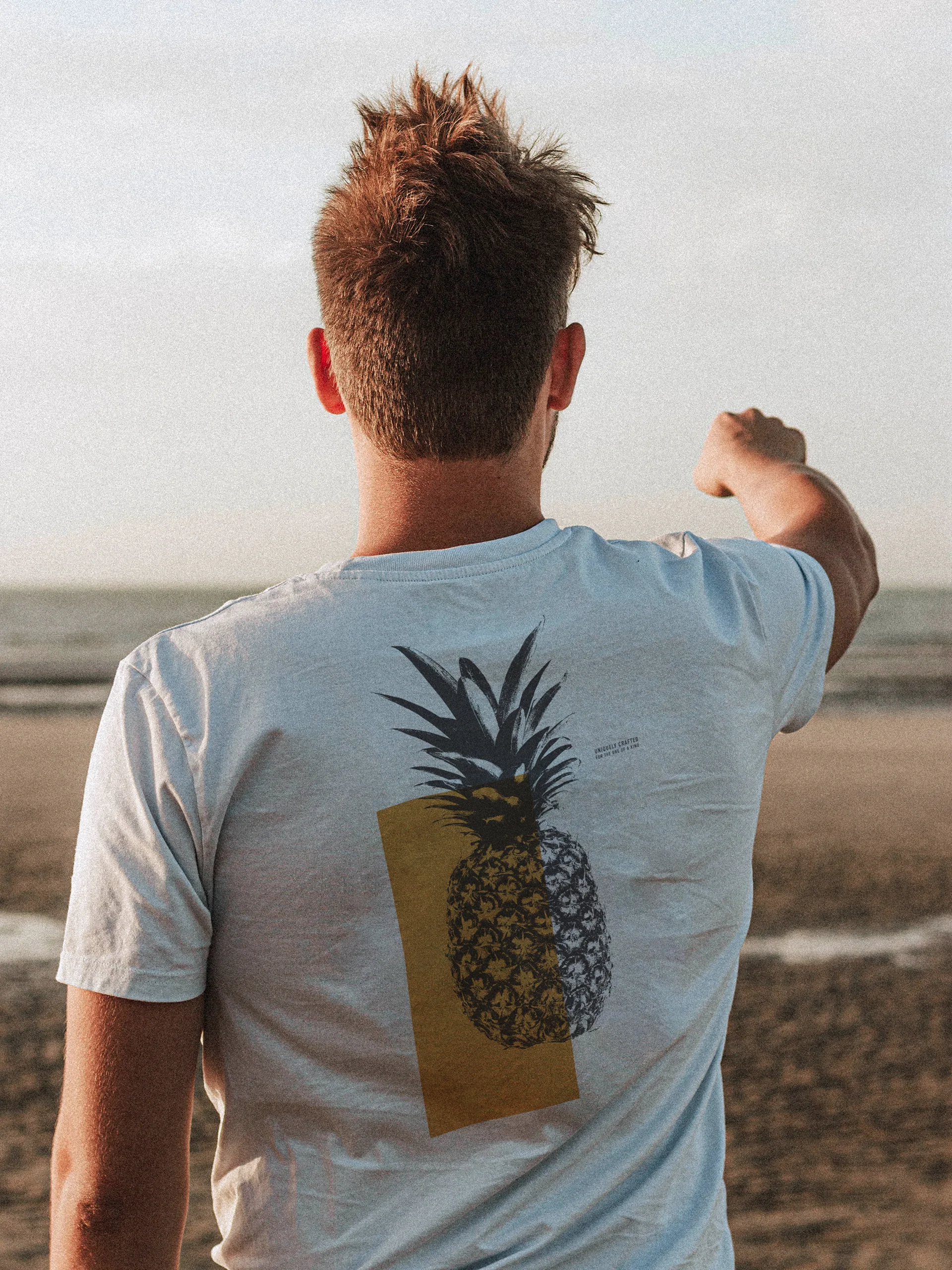 Man flexing his arm on a beach, wearing a light blue NIXED tee with a two-tone pineapple back print