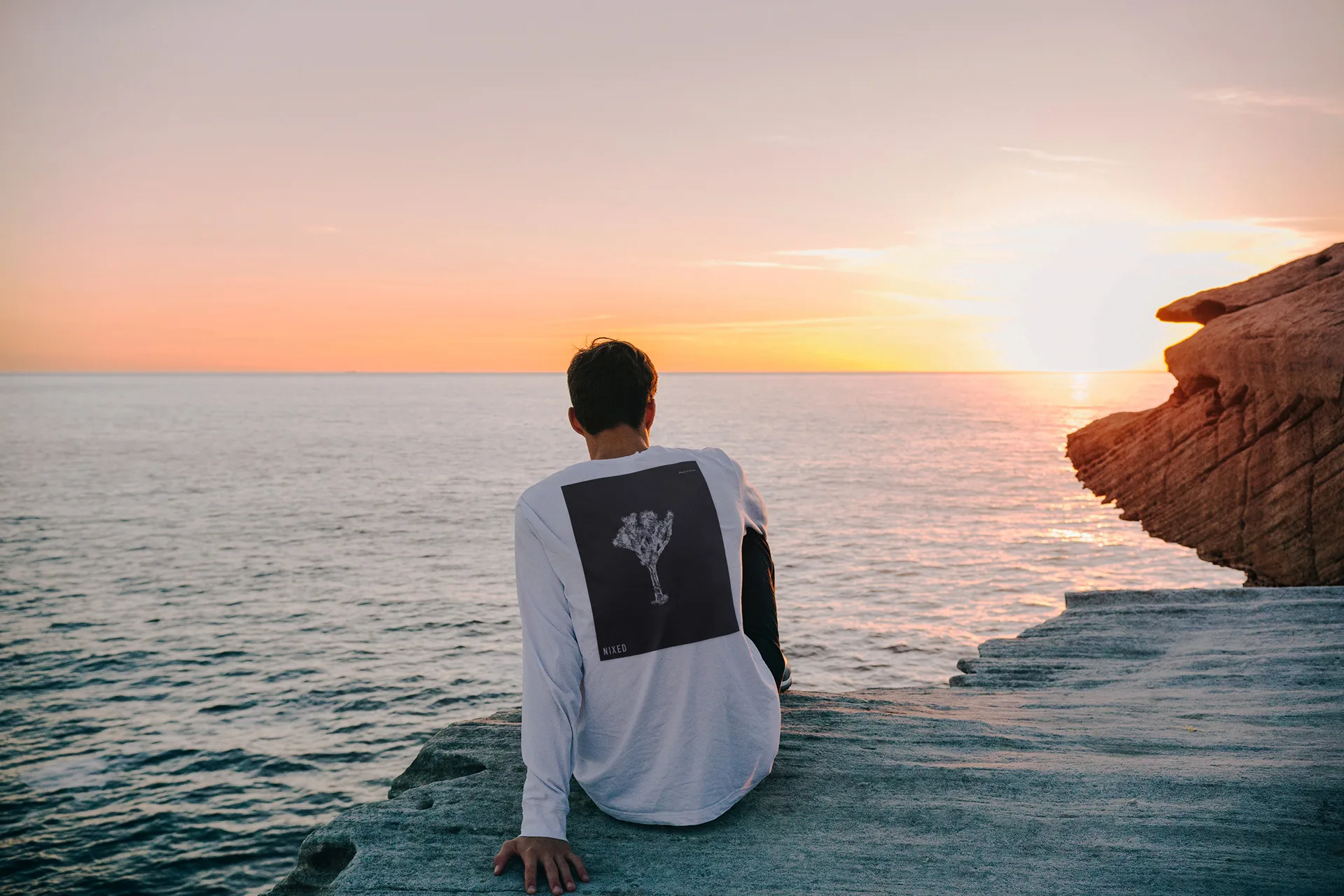 Man in a white long-sleeve NIXED tee with a small back graphic, sitting on coastal rocks at sunset