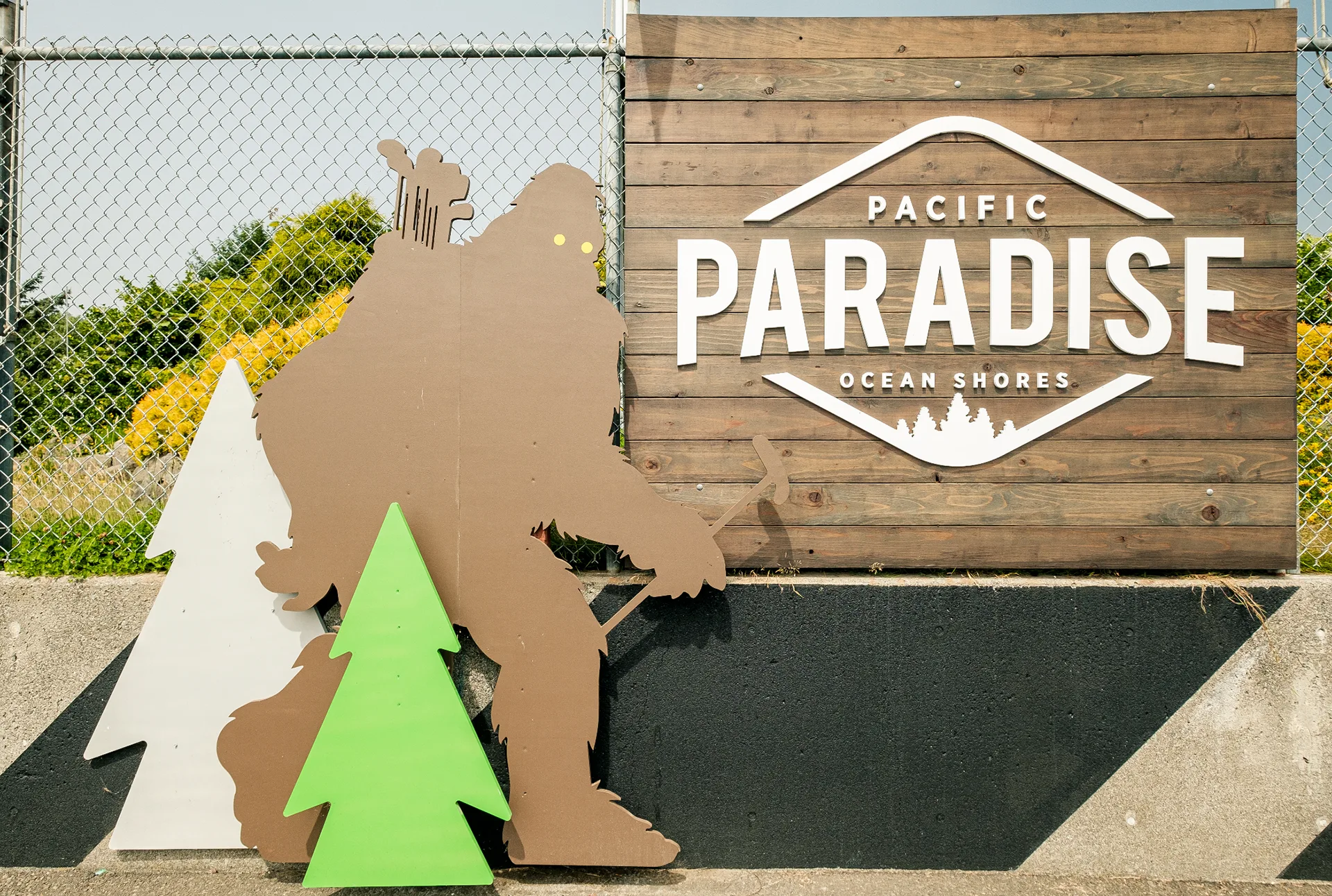 Pacific Paradise Ocean Shores entrance sign with bigfoot and pine tree cutouts against a chain-link fence and flowers