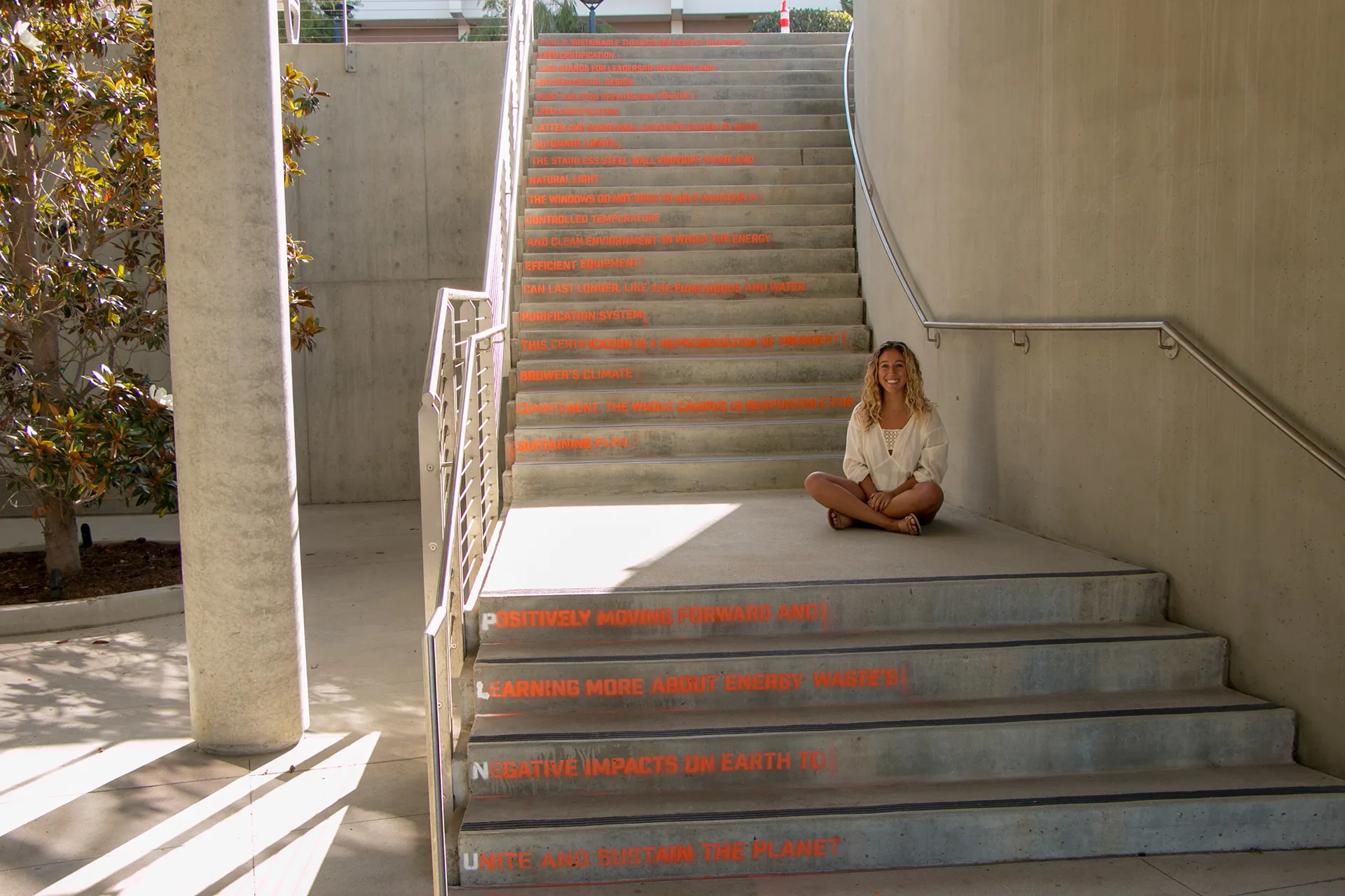 Woman sitting on outdoor stairs painted with orange sustainability facts about PLNU's environmental impact