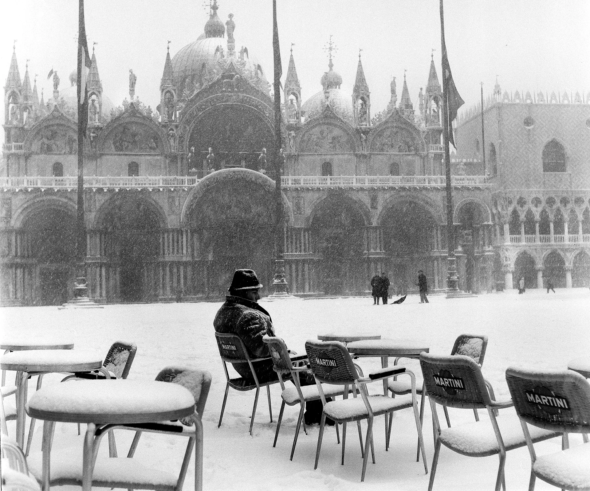 Black-and-white photo of St. Mark's Square in Venice during snowfall, with a man seated alone among empty café chairs