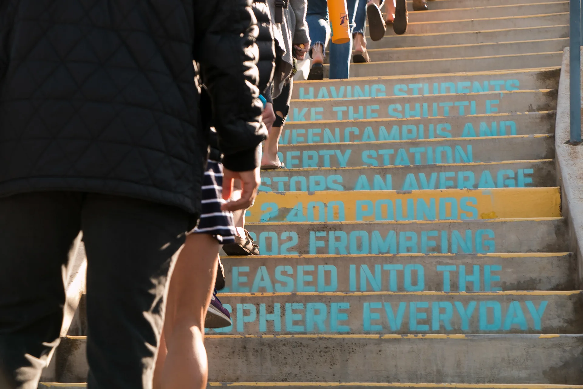Students walking down stairs stenciled with teal text about shuttle ridership reducing campus carbon emissions