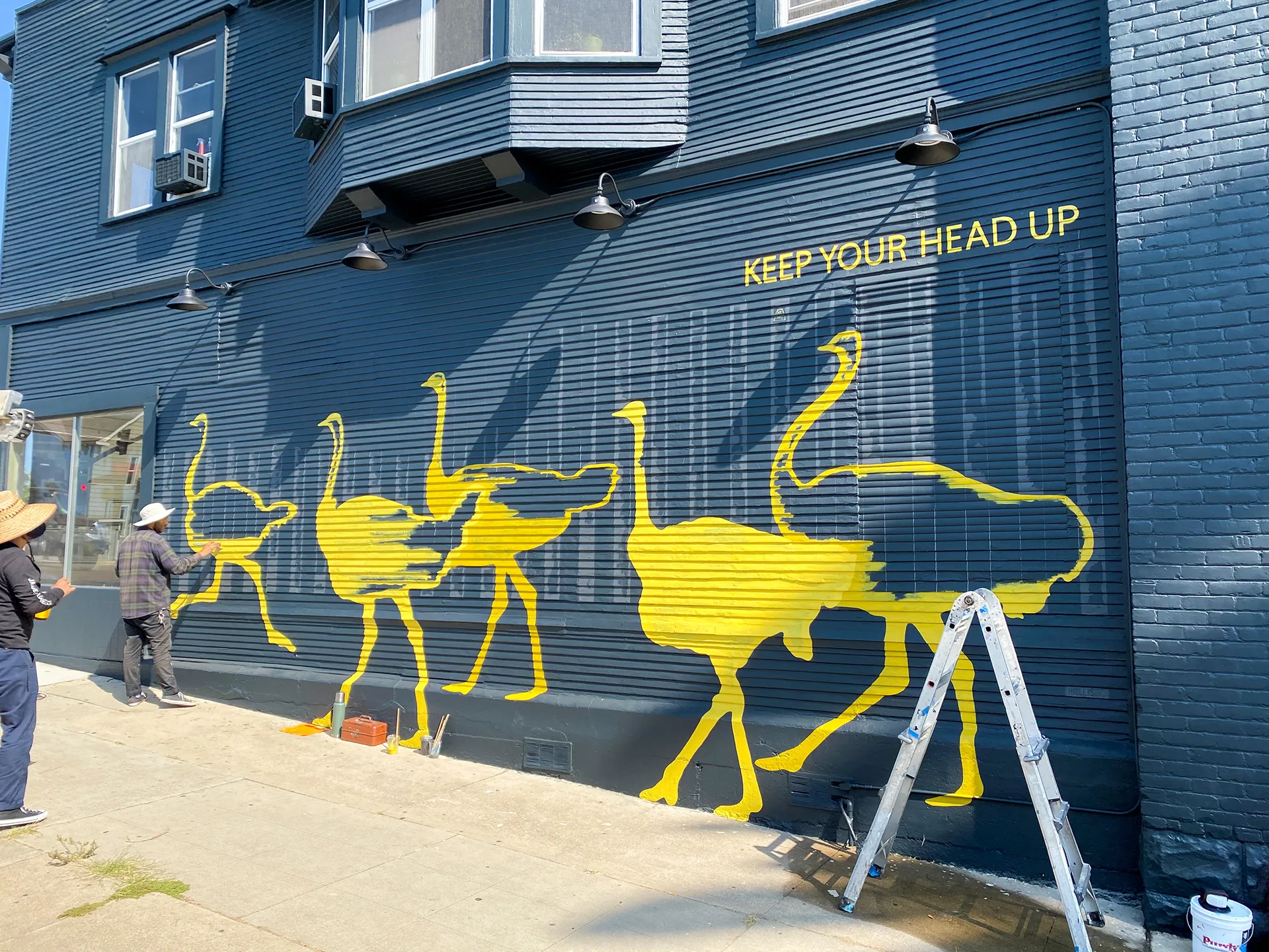 Ostrich mural in progress on the dark storefront wall, with ladders and supplies and two people reviewing the work
