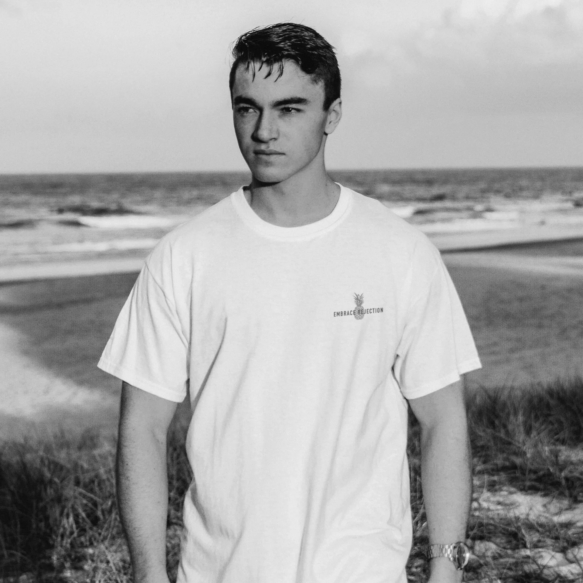 Man on the beach in a white NIXED tee with a small Embrace Rejection pineapple graphic, black-and-white photo