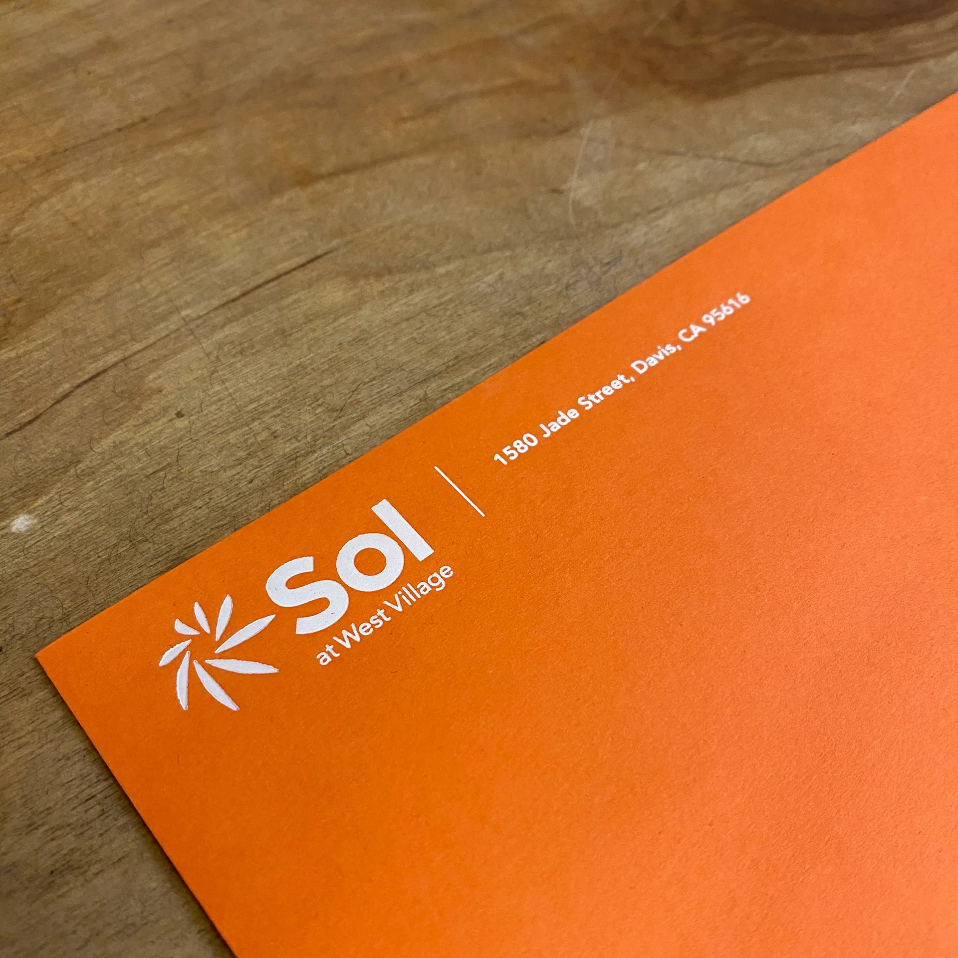 Close-up of an orange Sol at West Village envelope with the sunburst logo and address on a wood table