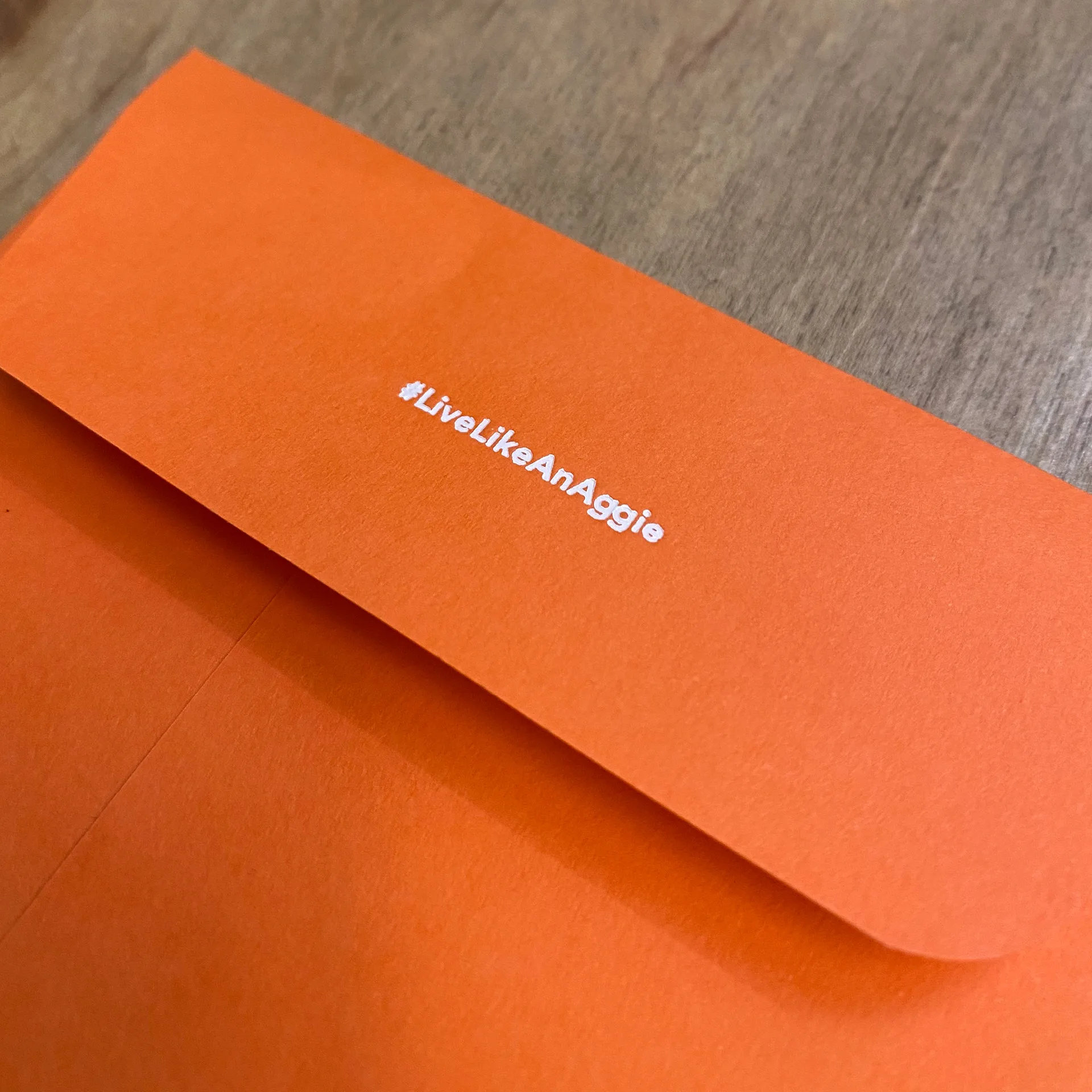 Close-up of an orange envelope flap printed with the hashtag LiveLikeAnAggie, resting on a wood table