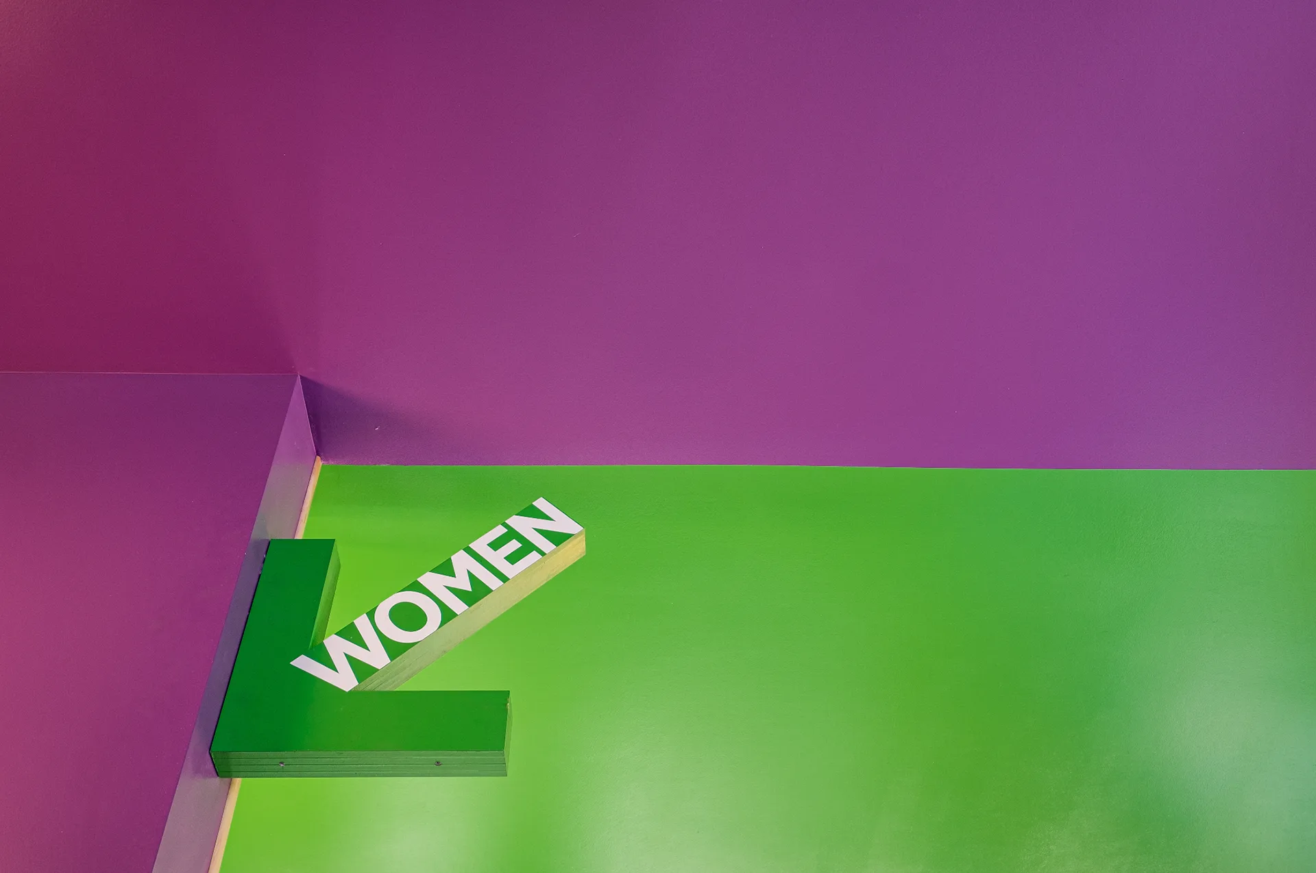 Green dimensional WOMEN arrow sign mounted where a purple wall meets a green ceiling