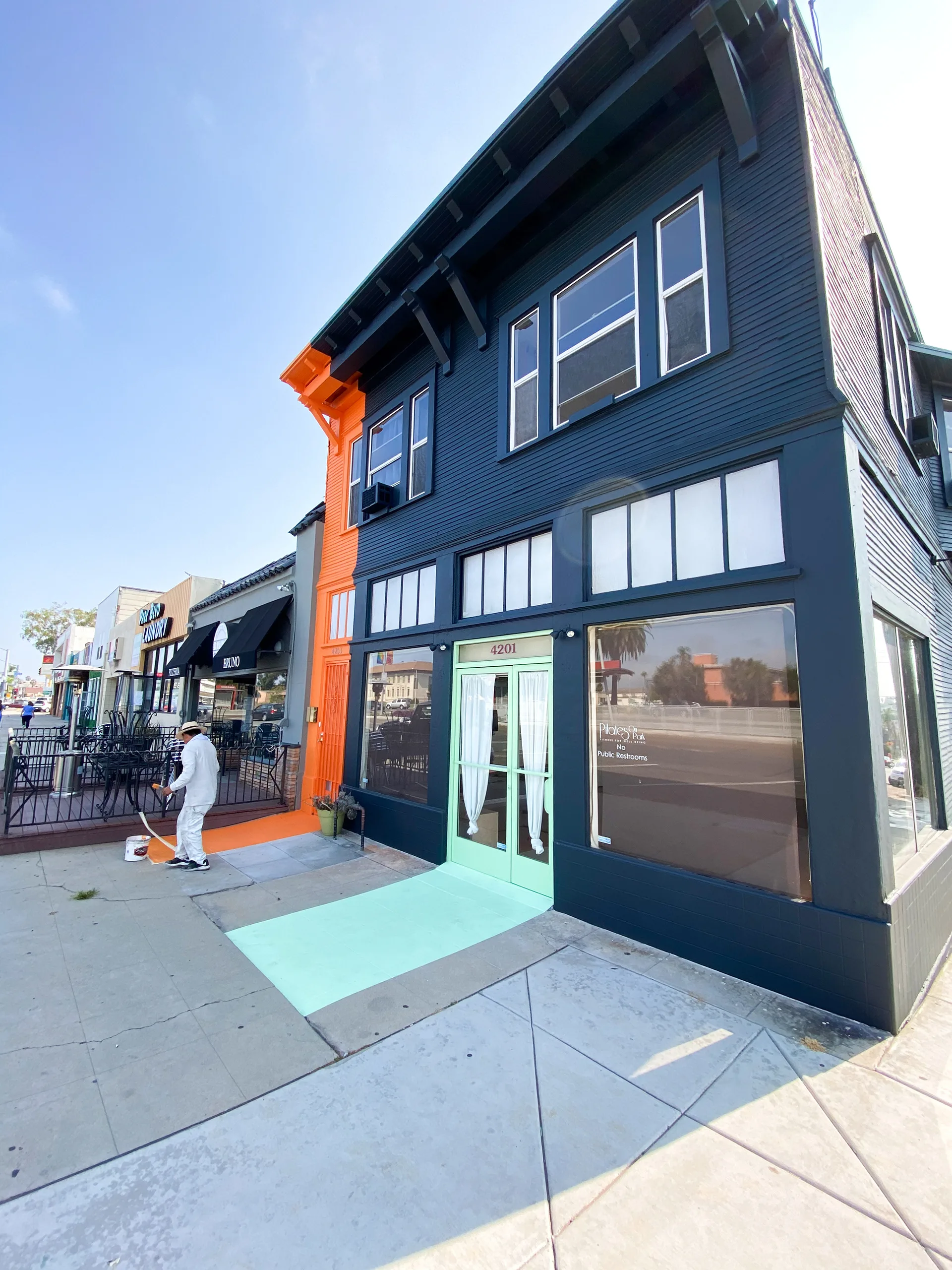 Wide street view of the finished orange-and-navy corner storefront with a mint green sidewalk path and a painter at work