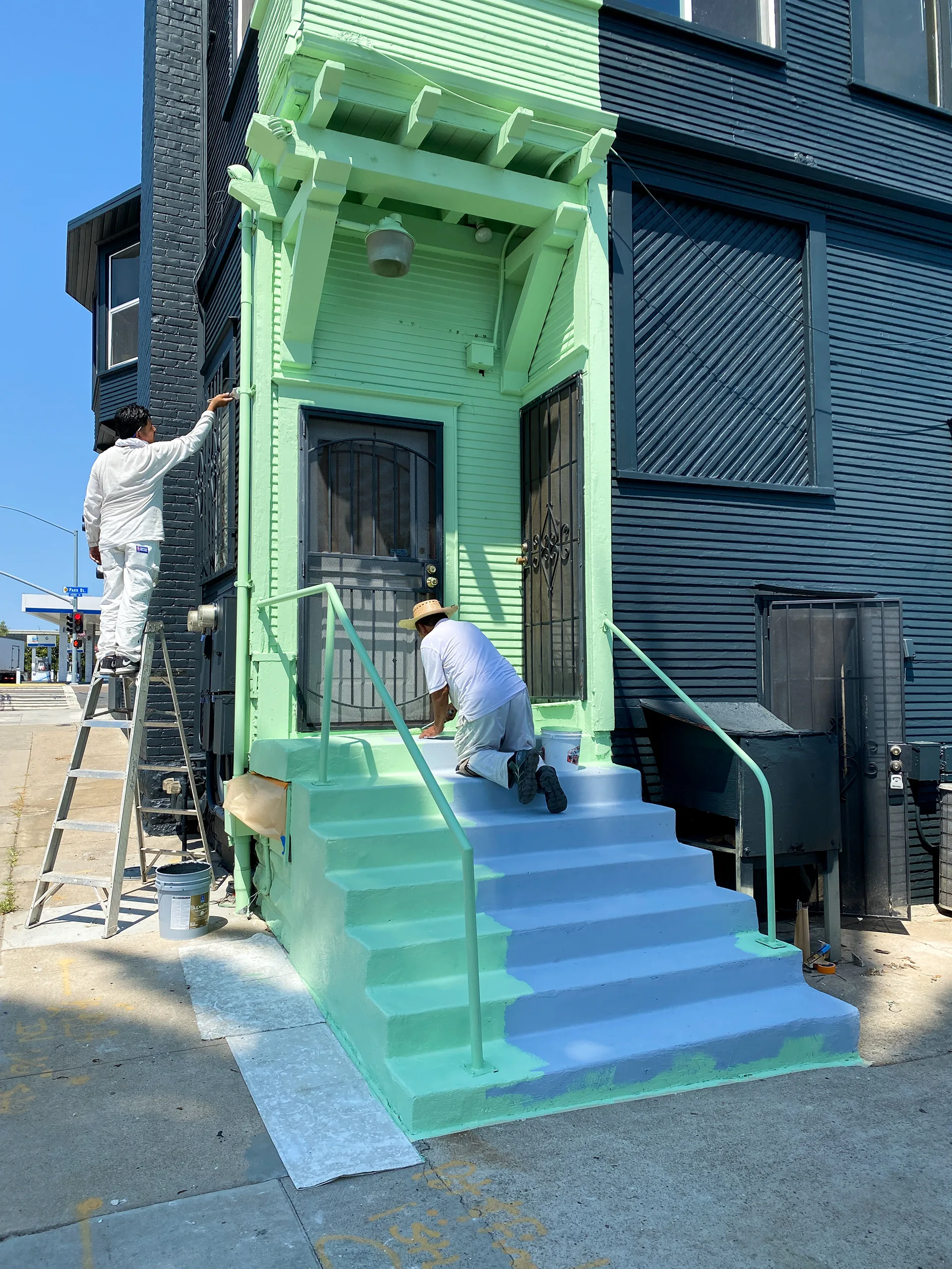 Two painters at work, one on a ladder finishing the green porch trim, one painting the entry stairs blue