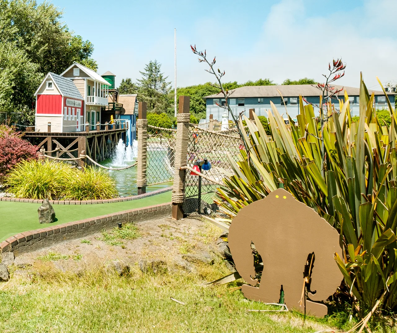Mini golf course green with lighthouse-style village buildings, a water feature, and a bigfoot cutout in tropical foliage