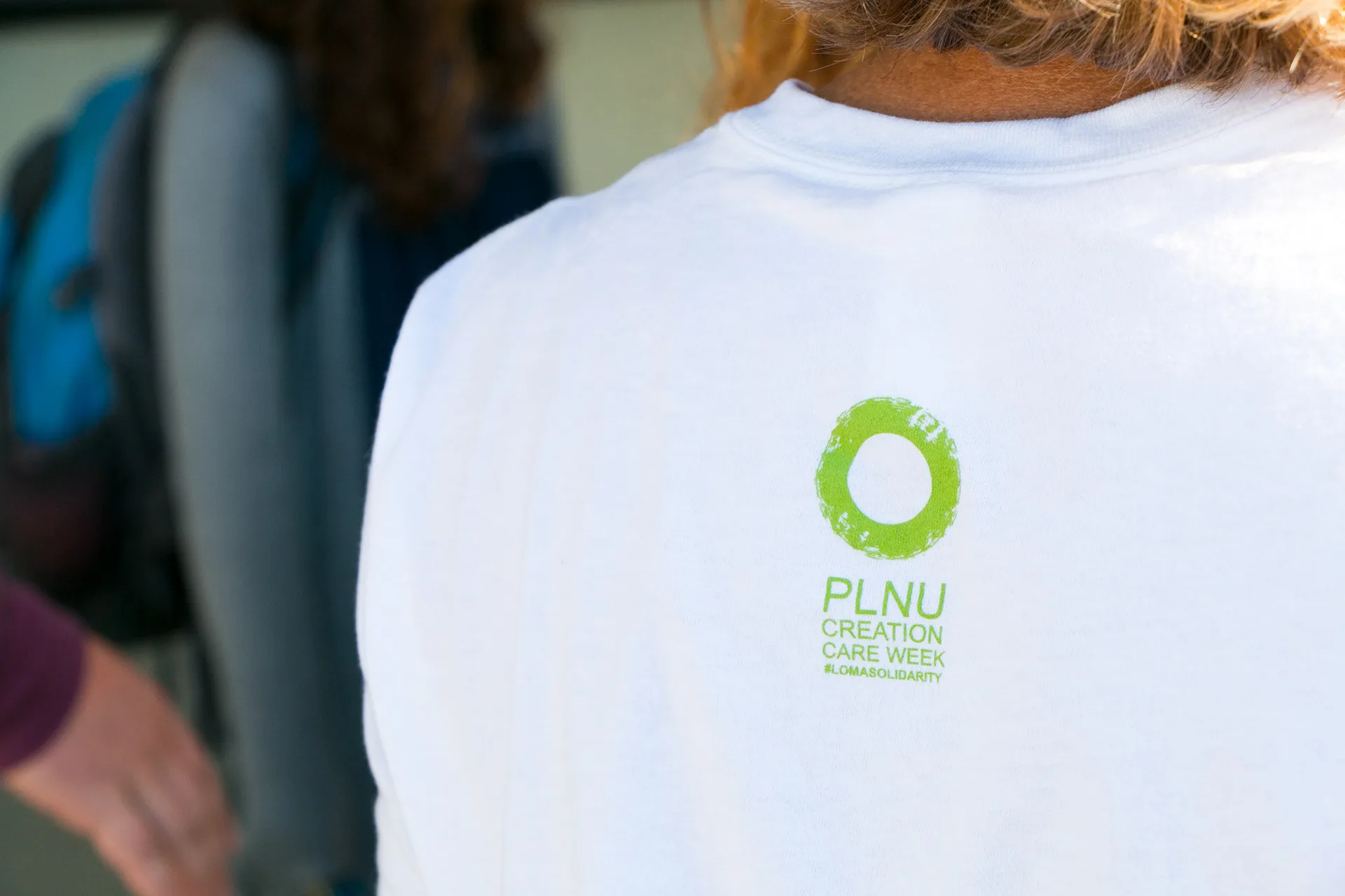 Close-up of a person's back wearing a white PLNU Creation Care Week t-shirt with a green circular logo