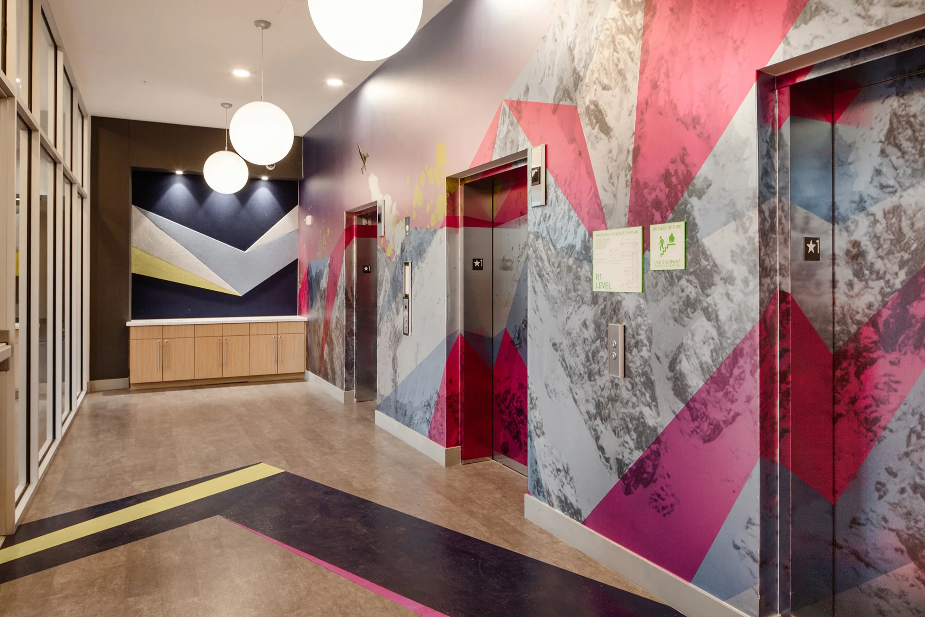 Elevator bank wrapped in a bold red, pink, and gray geometric mural with matching floor accent stripes