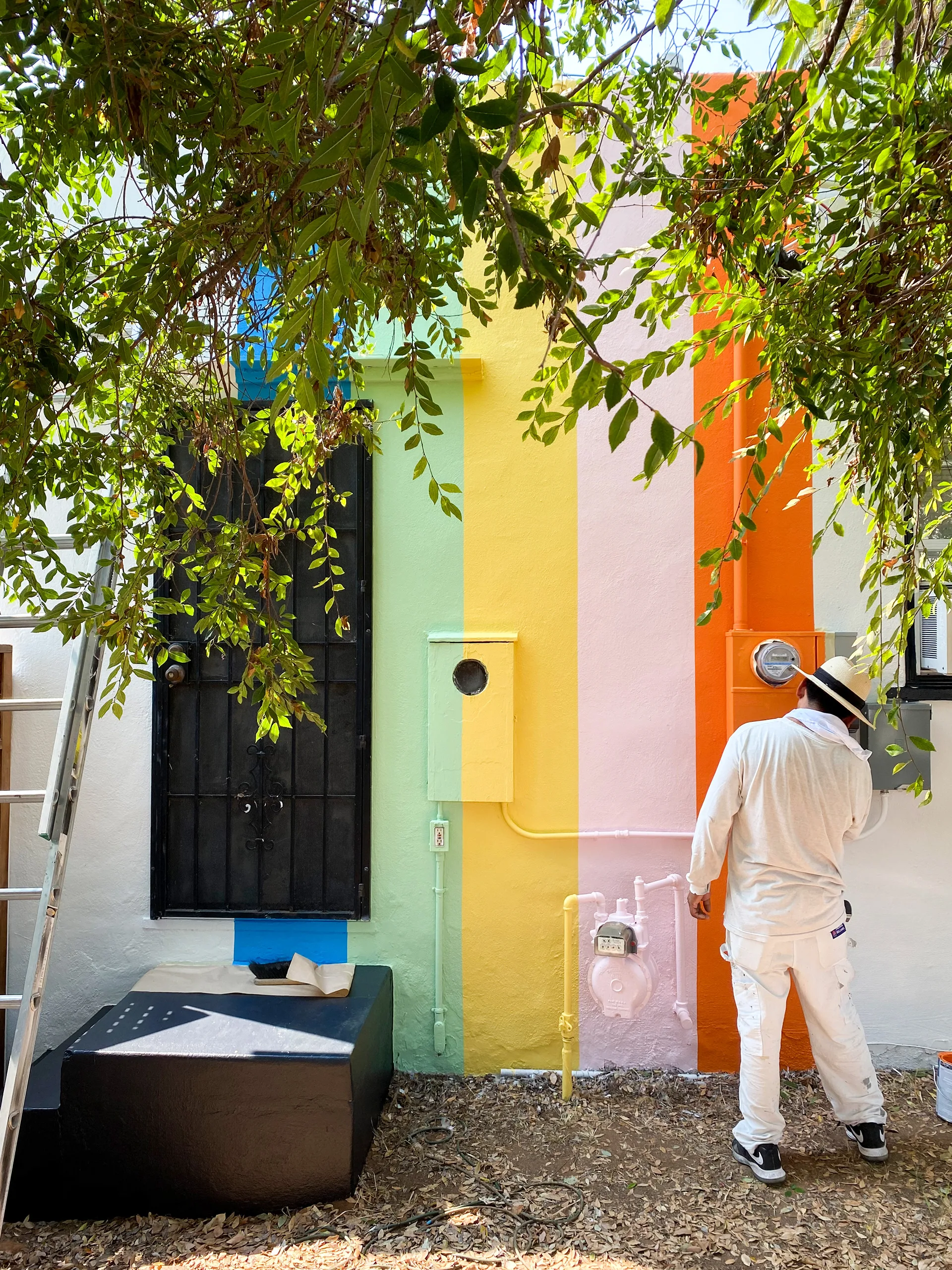 Painter in white coveralls working on the rainbow-striped wall beneath overhanging tree branches, ladder leaning against the wall