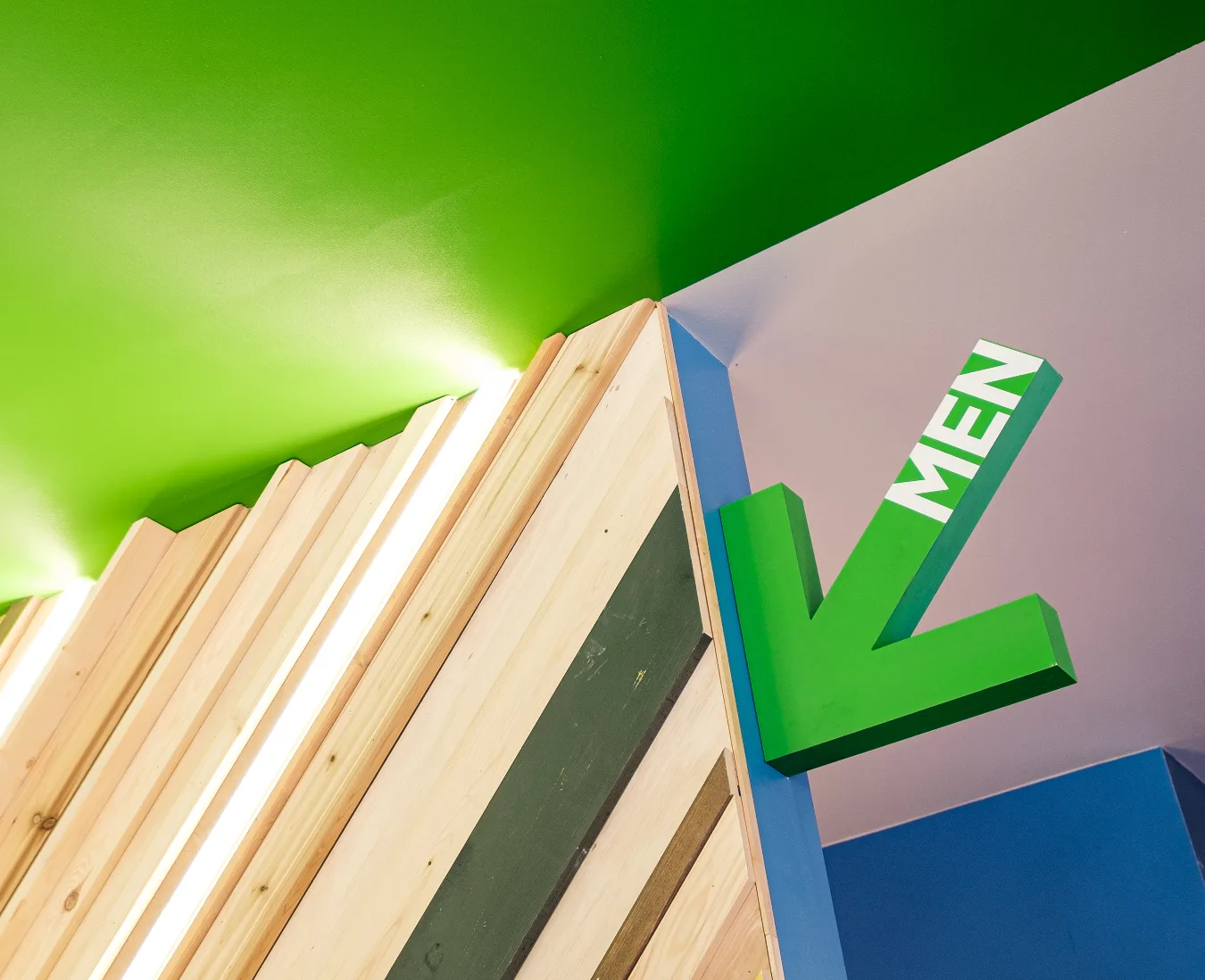 Green dimensional MEN arrow sign mounted on a corner above a backlit wood slat accent wall