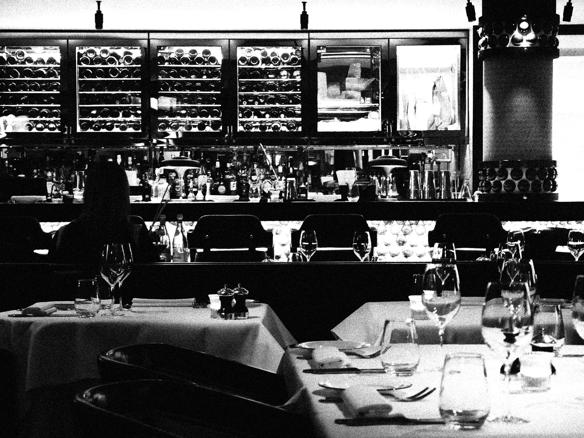 Black-and-white photo of a dim restaurant bar and dining area with wine racks and set tables