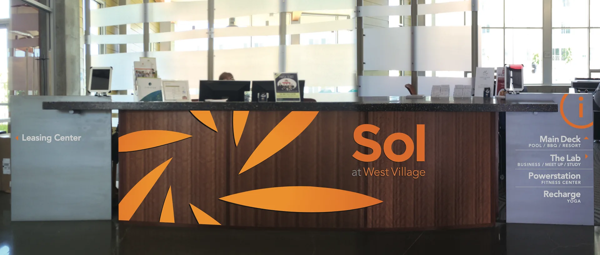 Sol at West Village leasing center desk with a wood panel sunburst logo and an amenities directory sign beside it