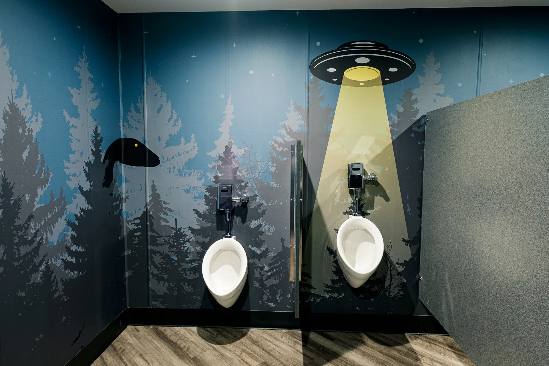 Men's restroom urinals set against a night forest mural with a bigfoot silhouette and glowing UFO graphic
