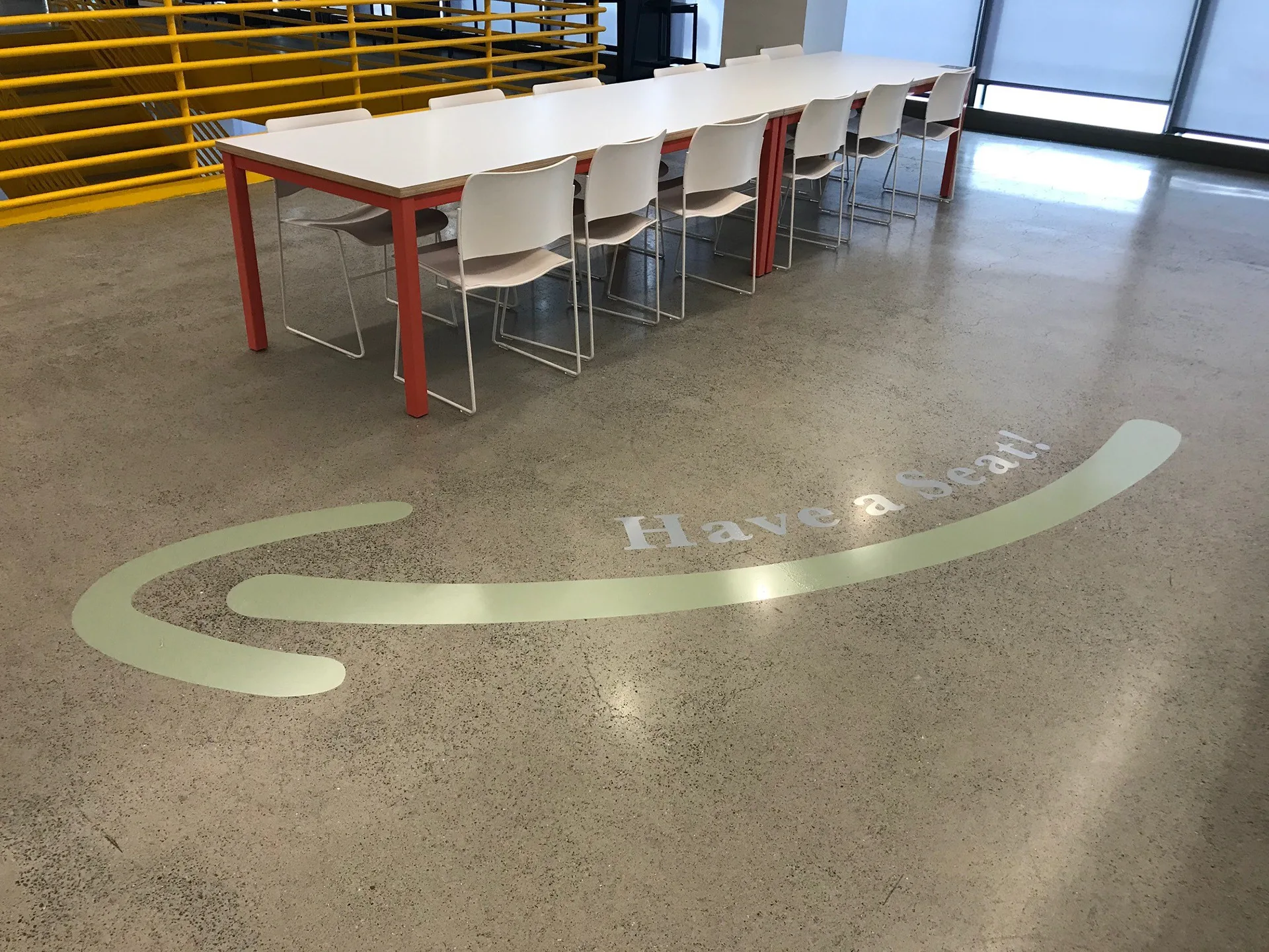 Have a Seat floor graphic beside a long communal dining table with red-framed chairs