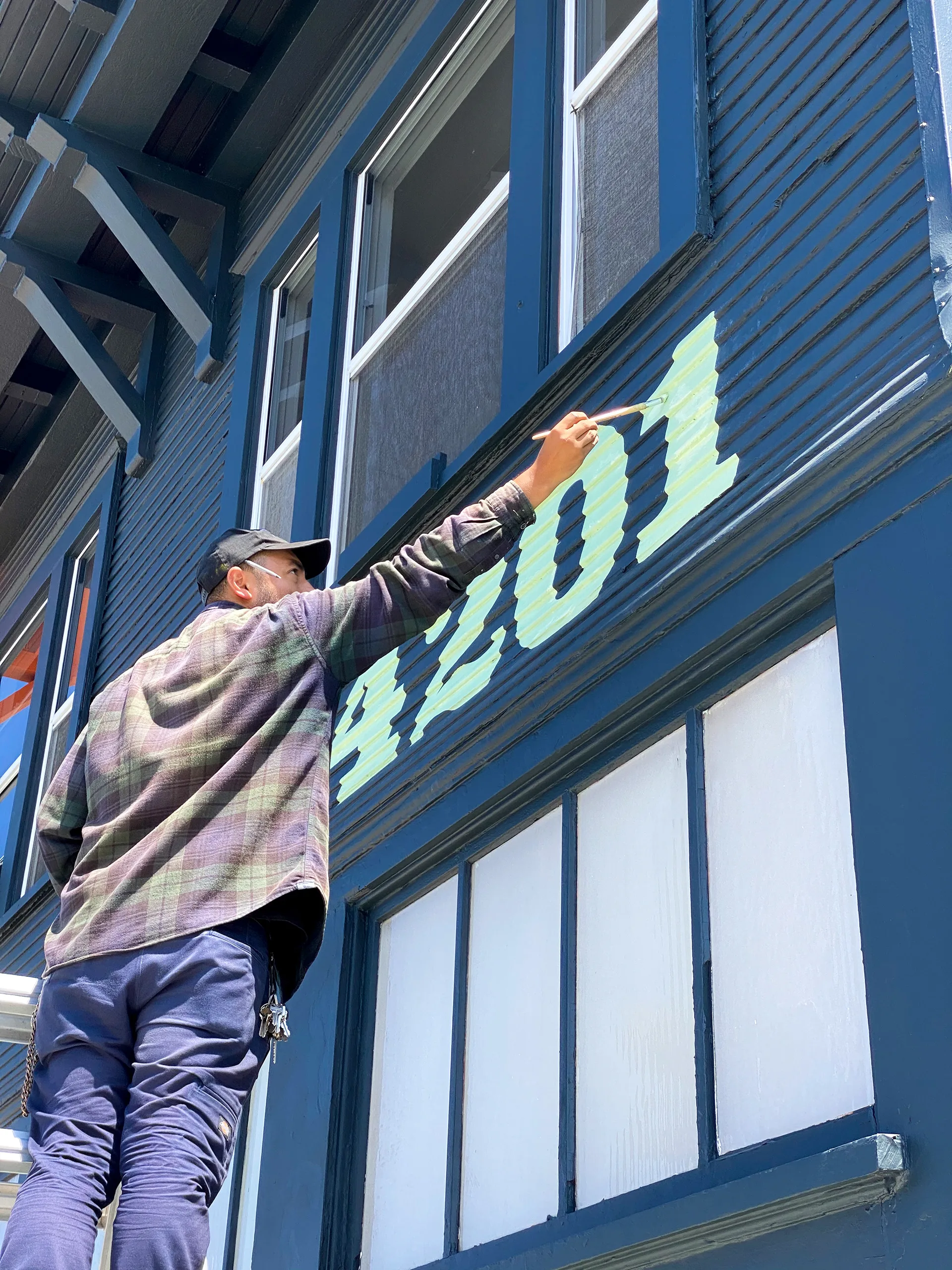 Painter on a ladder hand-painting the mint green 4201 address numerals onto the navy building siding