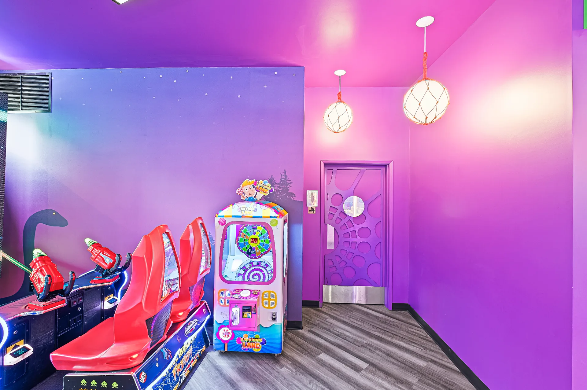 Purple-lit arcade room with racing game seats, a prize claw machine, and a spider-web patterned door