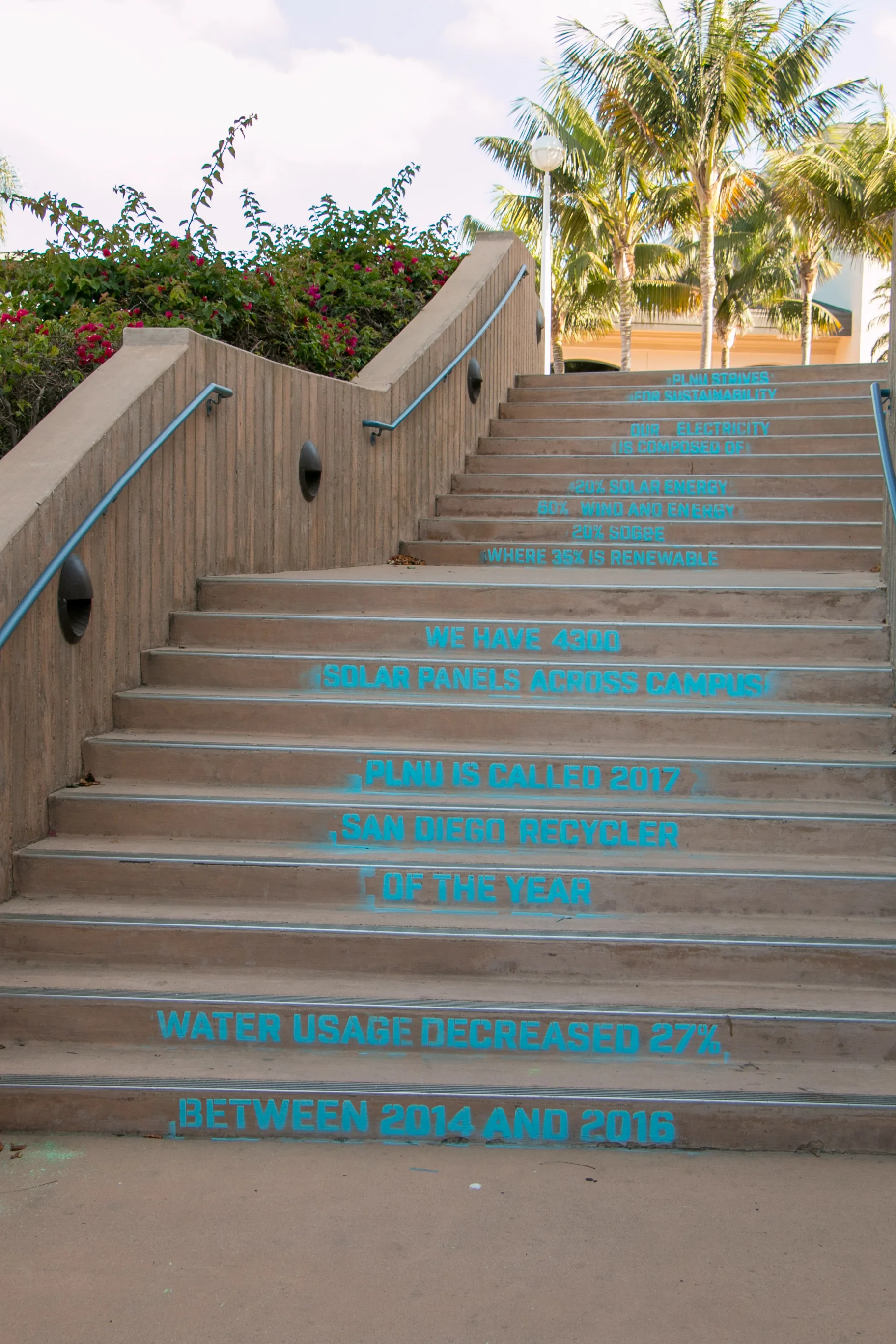 Outdoor staircase stenciled in teal with statistics on solar panels, recycling awards, and water usage reduction at PLNU