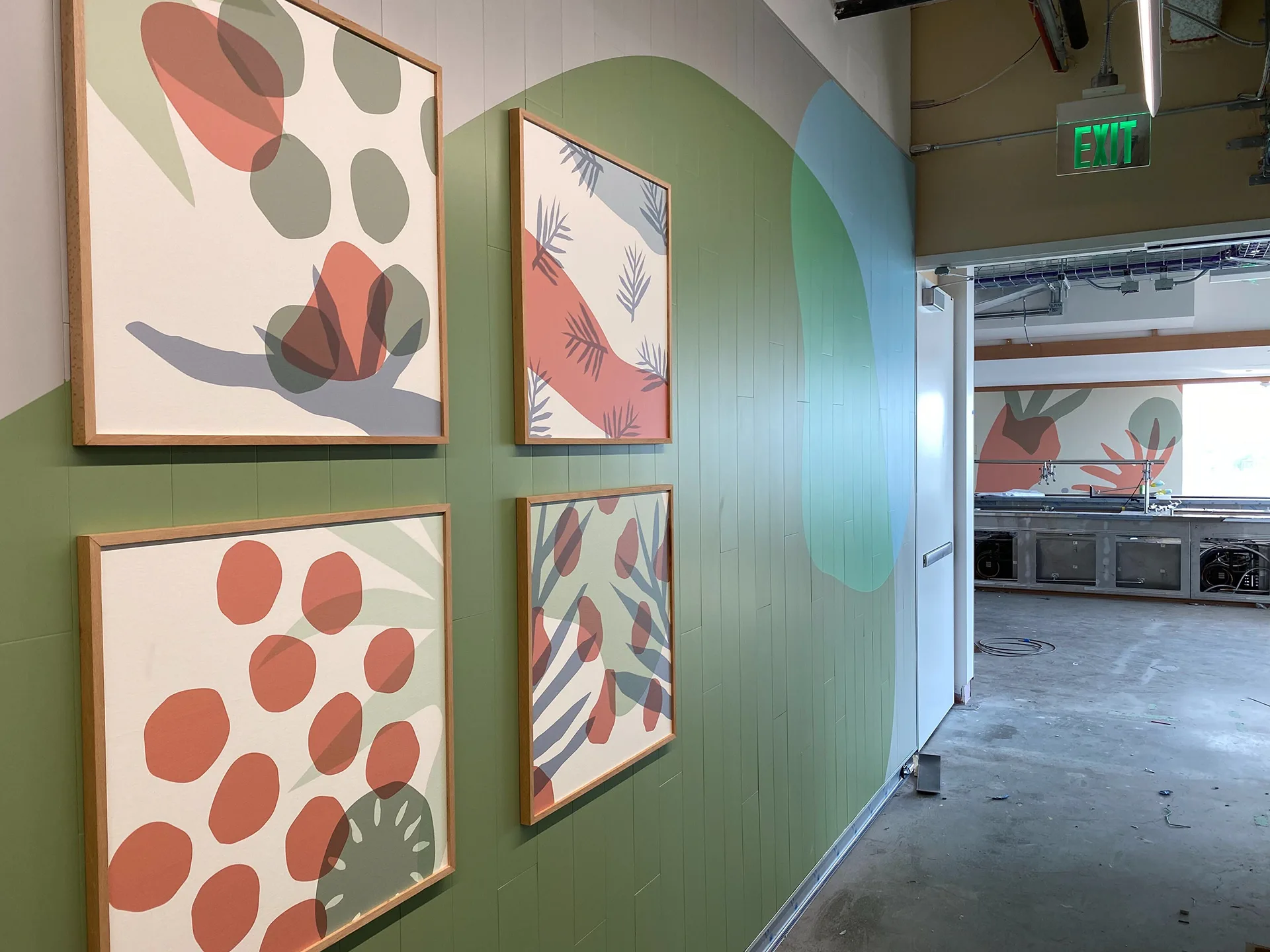 Four framed produce-pattern art prints on a green mural wall near the kitchen entrance and exit sign