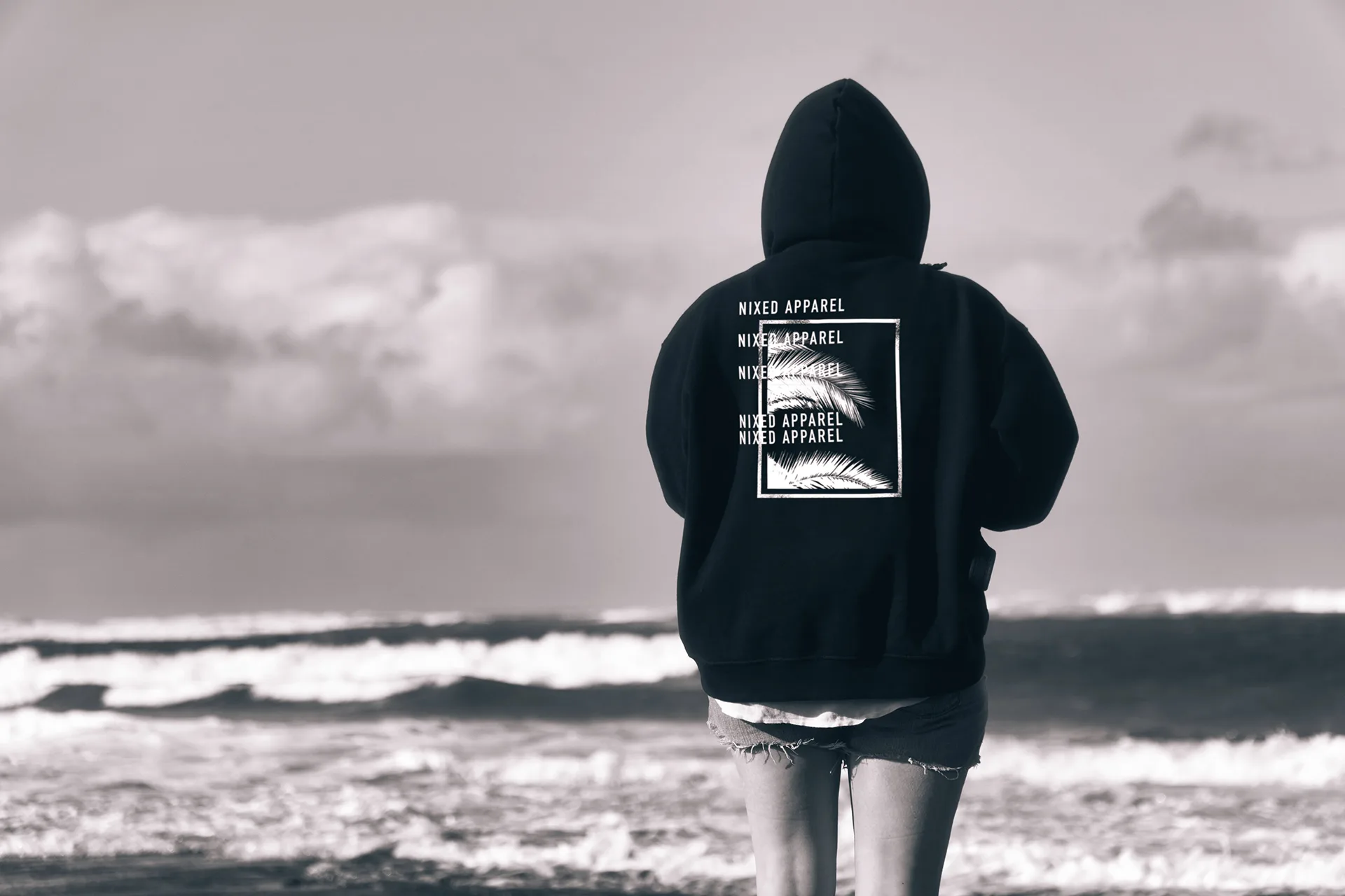 Woman's back in a NIXED Apparel hoodie with repeated wordmark and palm print, standing on a beach, black-and-white photo