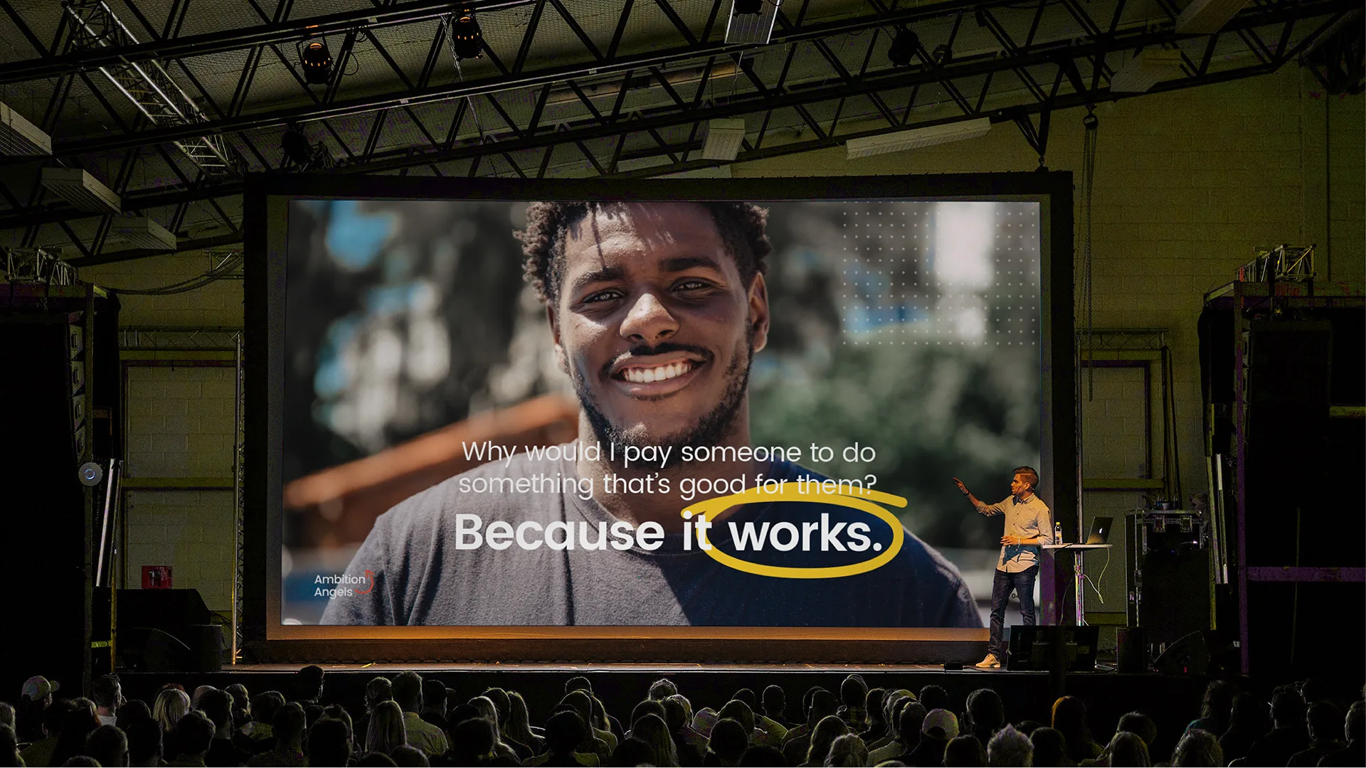 Conference stage with a large screen displaying a smiling man's portrait and the line because it works, speaker gesturing below