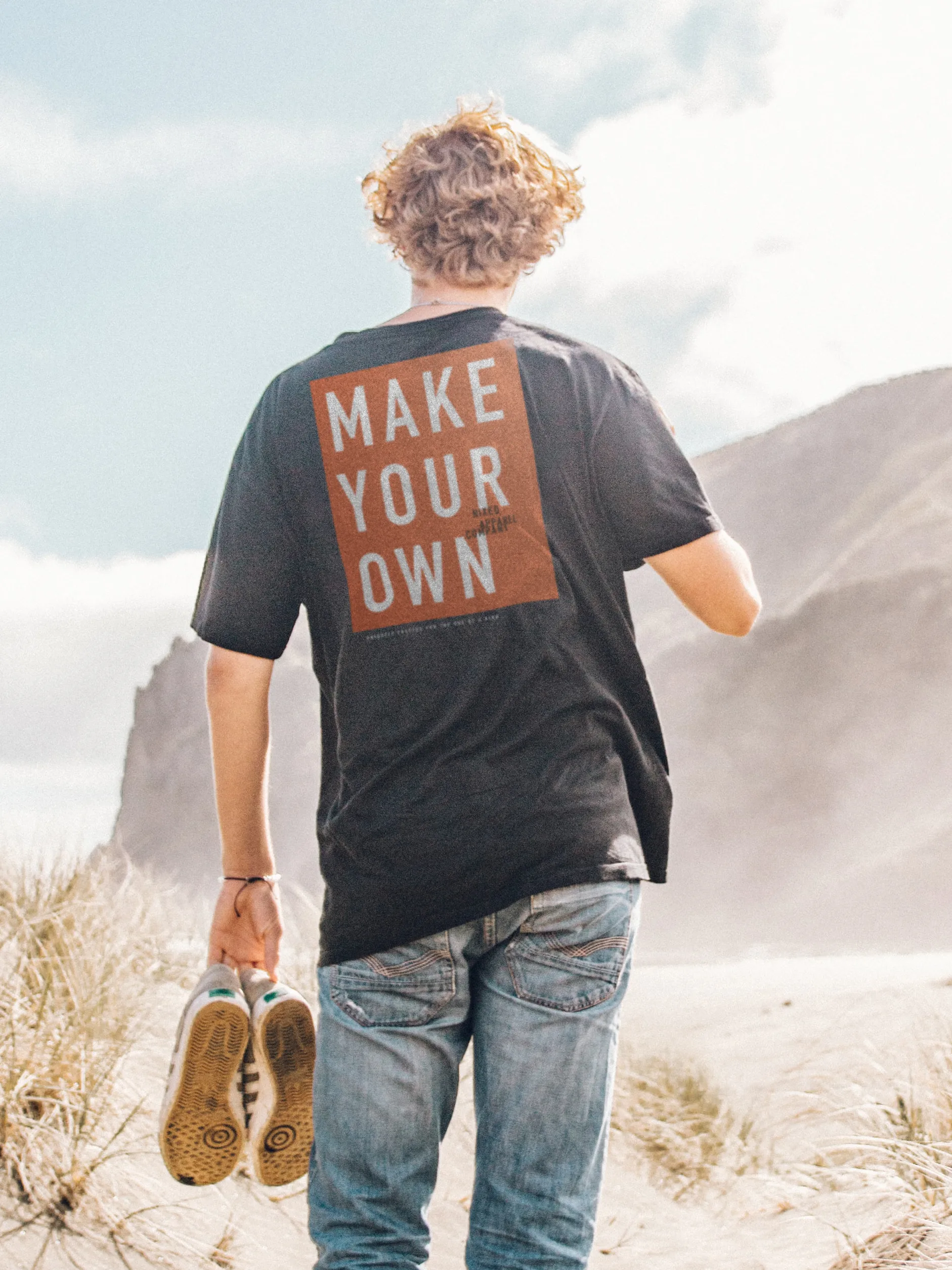 Man's back in a black NIXED tee reading Make Your Own, standing on sandy dunes holding his shoes