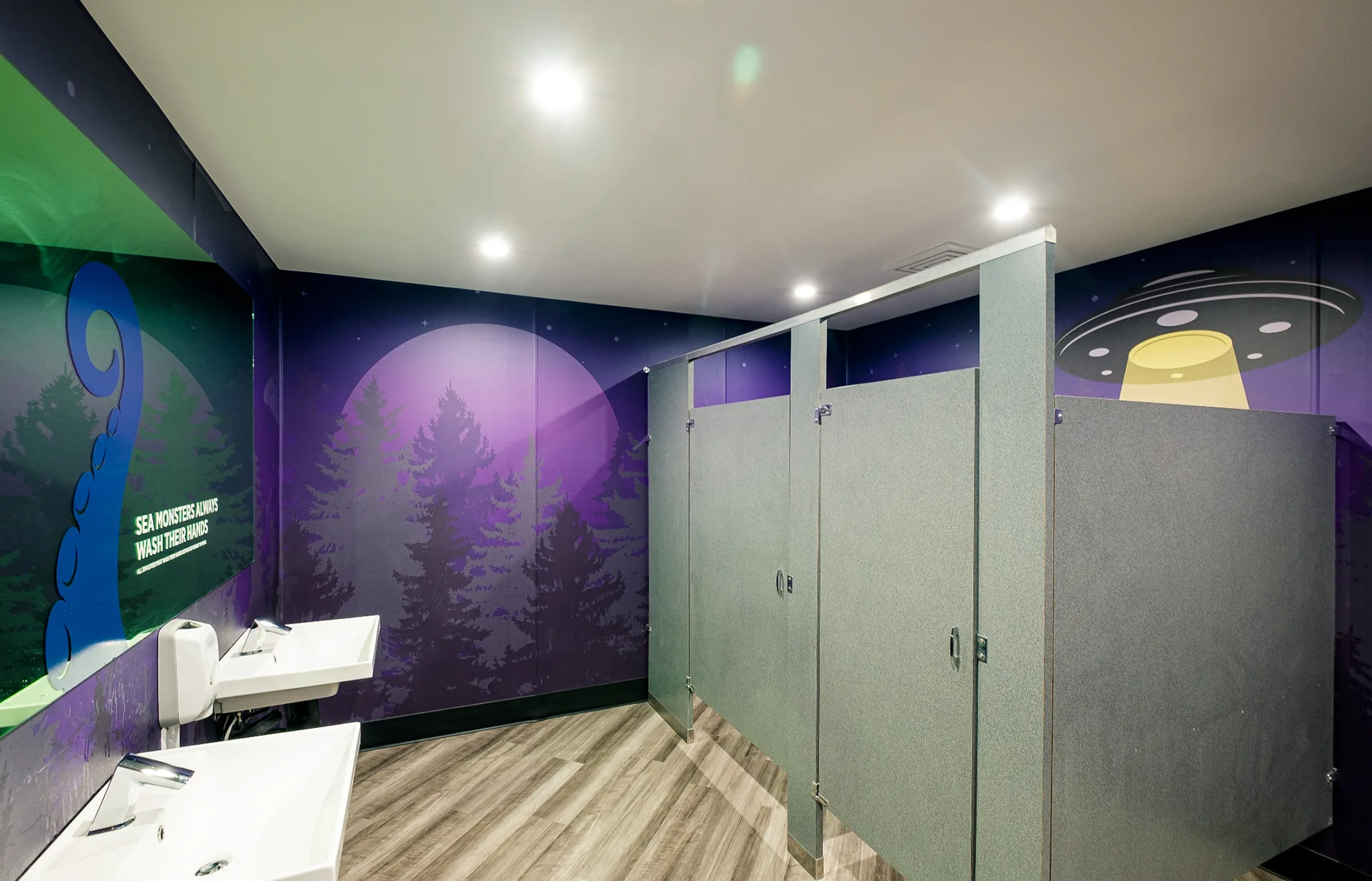 Restroom stalls with a purple moonlit forest mural reading 'sea monsters always wash their hands' and a UFO graphic