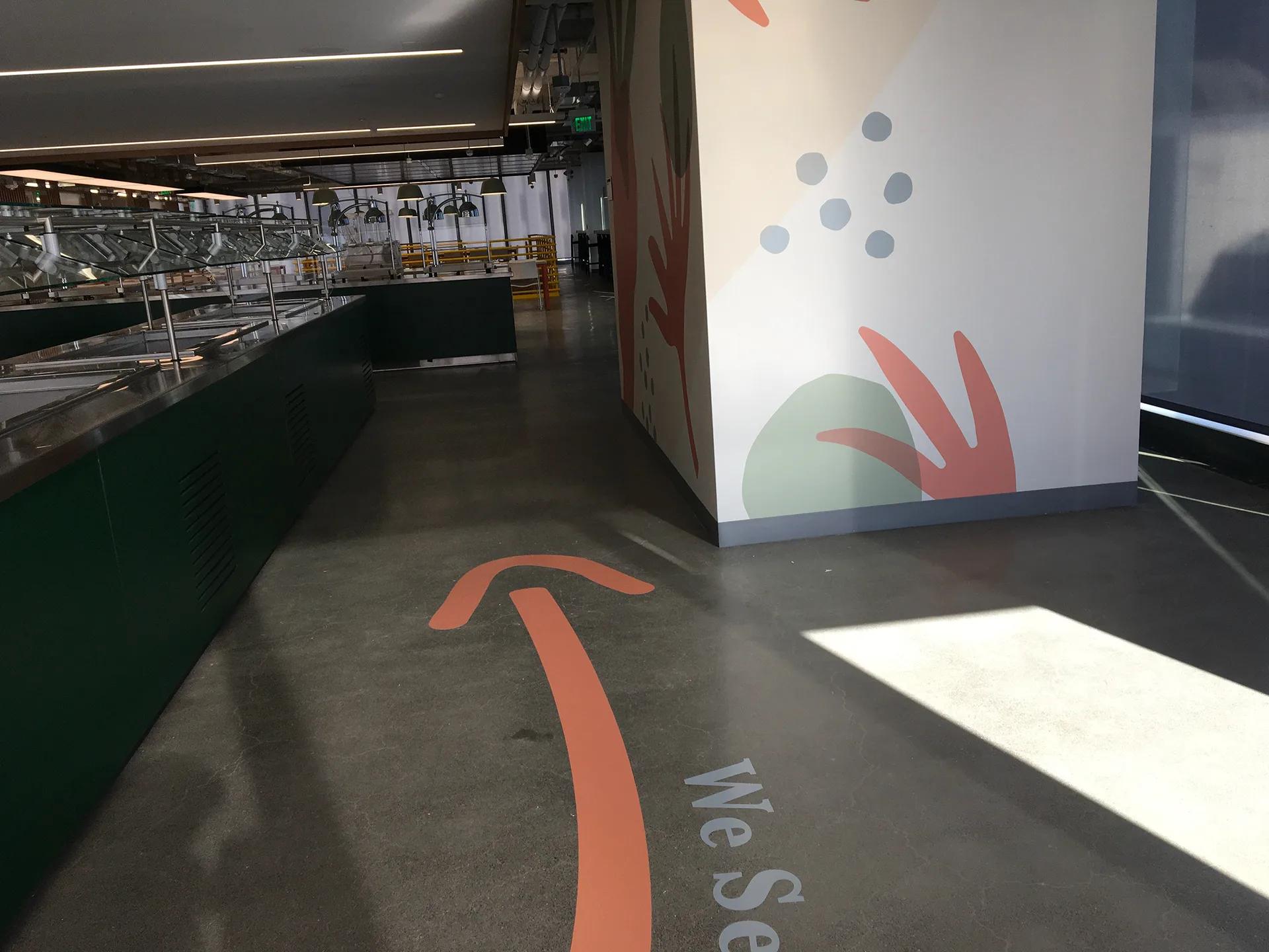 We Serve You floor arrow leading past the serving line toward a painted botanical wall mural