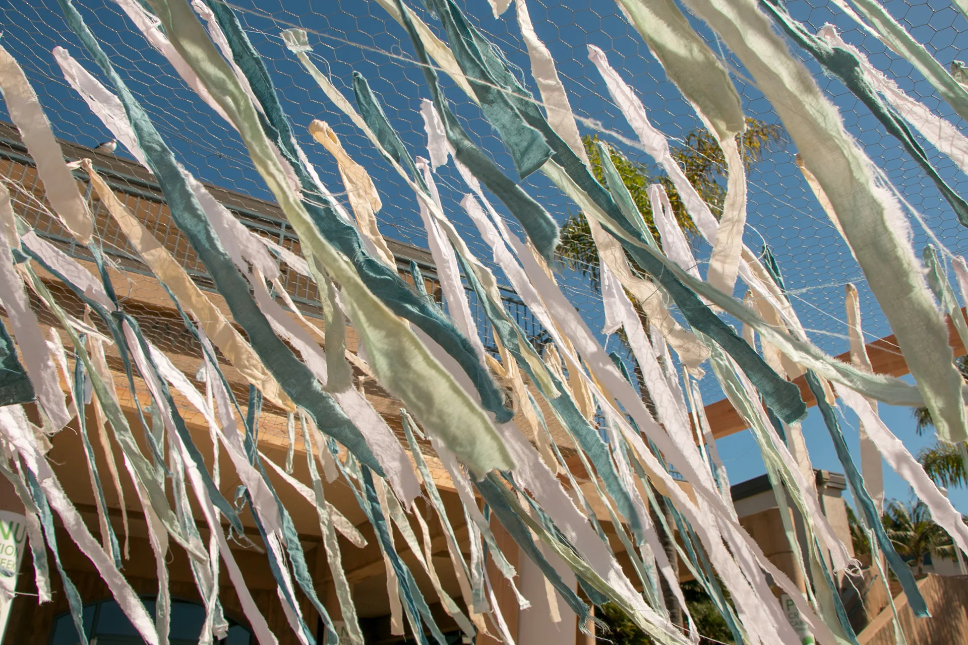 Close-up of pastel fabric ribbon strips strung overhead for the pledge installation, blue sky and rooftop behind
