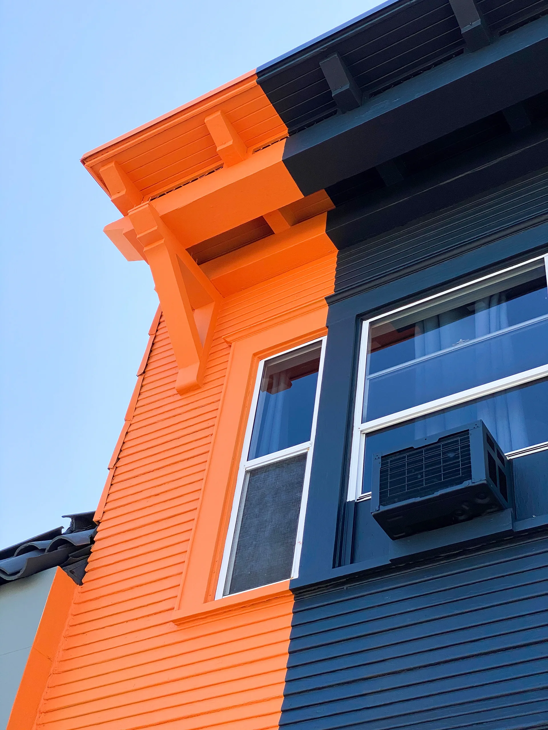 Close-up of the roofline cornice where the building's orange and navy paint blocks meet, with a window AC unit